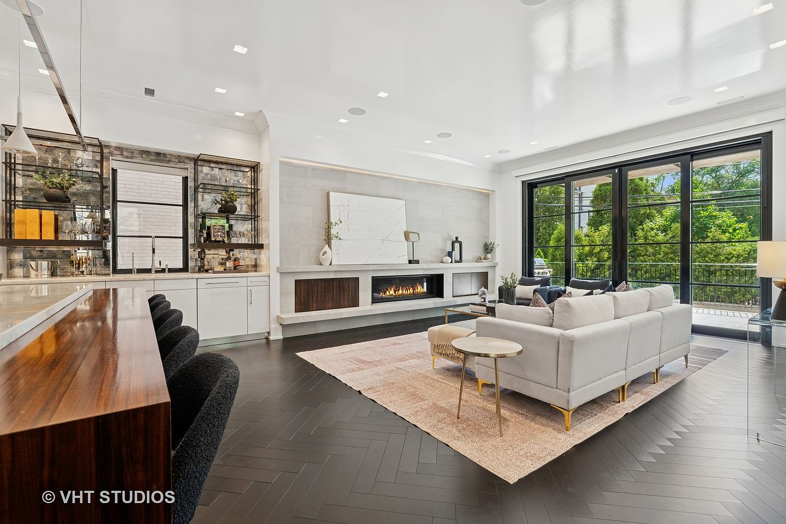 This is a bright and modern living room featuring dark herringbone wood floors, a large area rug, and a contemporary fireplace with a marble surround. A wet bar is located on the left side of the room. The room has a large sliding glass door that leads to a balcony, providing ample natural light and a view of the outdoors. The perspective is from the front of the room, looking towards the fireplace and balcony.