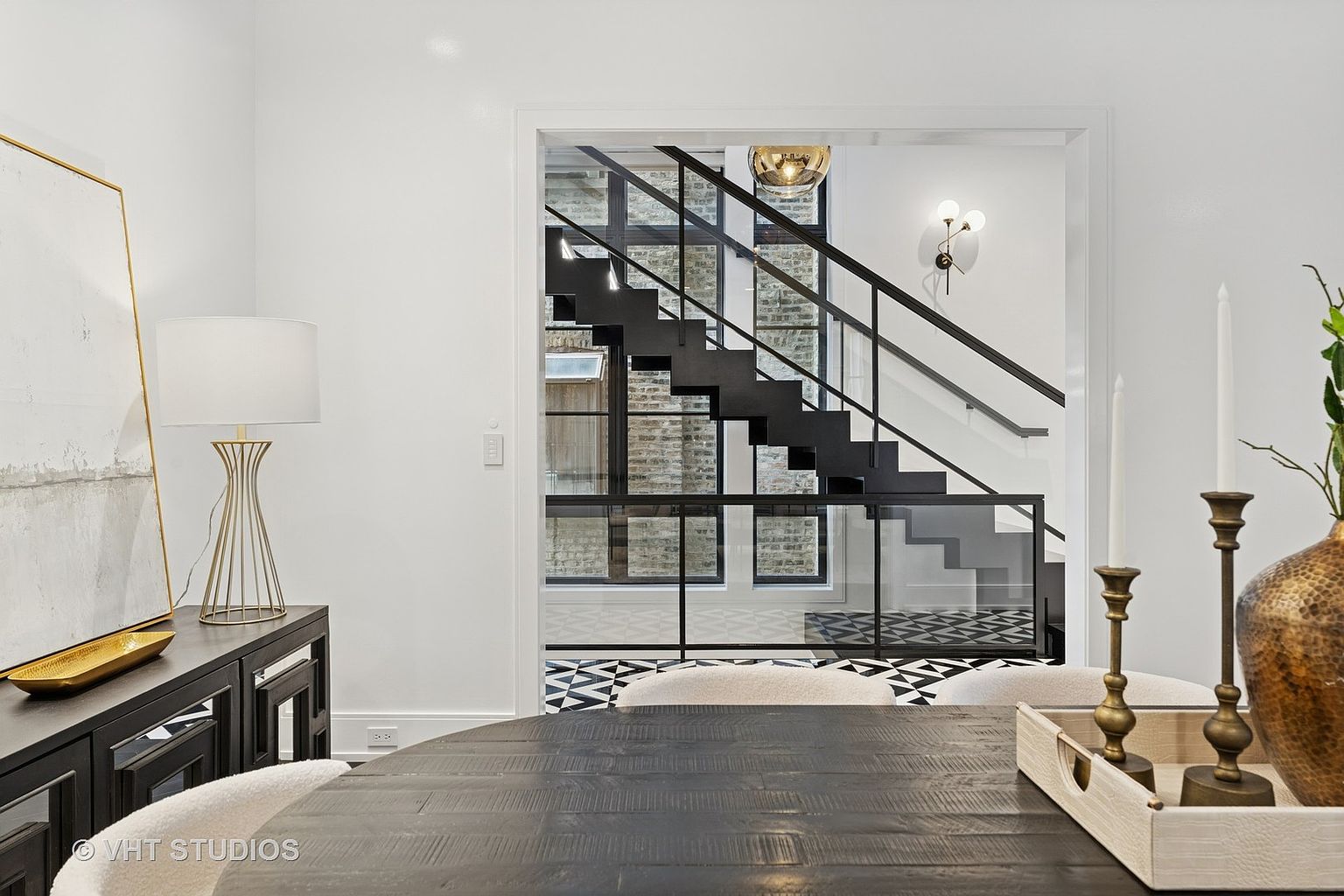 This interior shot showcases a dining room with a modern aesthetic. A dark, round wooden table takes center stage, complemented by light-colored chairs. The room opens to a staircase with black metal railings and glass panels, adding an architectural focal point. The flooring features a striking black and white geometric pattern, enhancing the contemporary feel of the space.