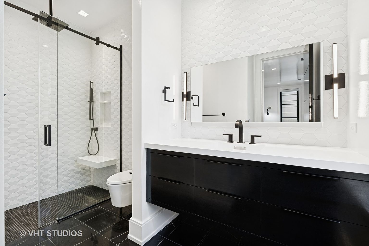 This is a modern primary bathroom featuring a glass-enclosed shower with black hardware and a built-in bench. The vanity has a sleek, black cabinet with a white countertop, complemented by a large mirror and modern light fixtures. The black tile flooring adds a touch of elegance and contrasts with the white walls and geometric tile backsplash.