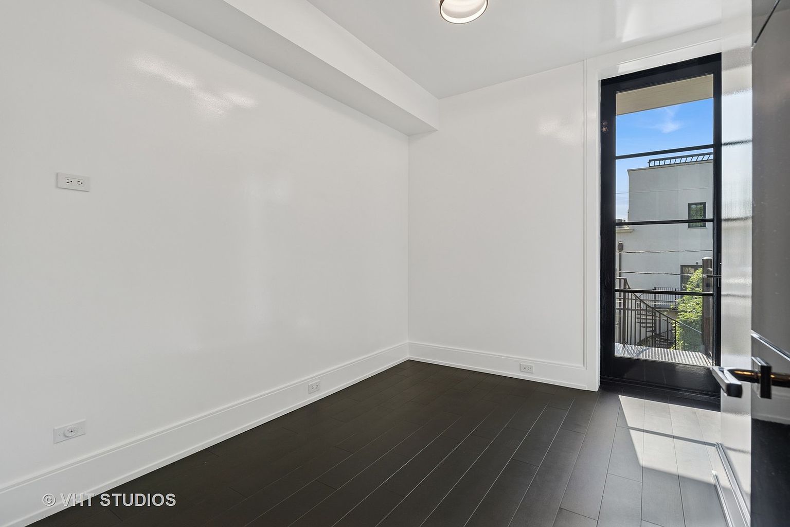 The image showcases a bright, minimalist bedroom with white walls and dark hardwood flooring. A black-framed glass door leads to an outdoor area, providing natural light and a view of the exterior. The room is clean and uncluttered, emphasizing a sense of spaciousness and modern design.