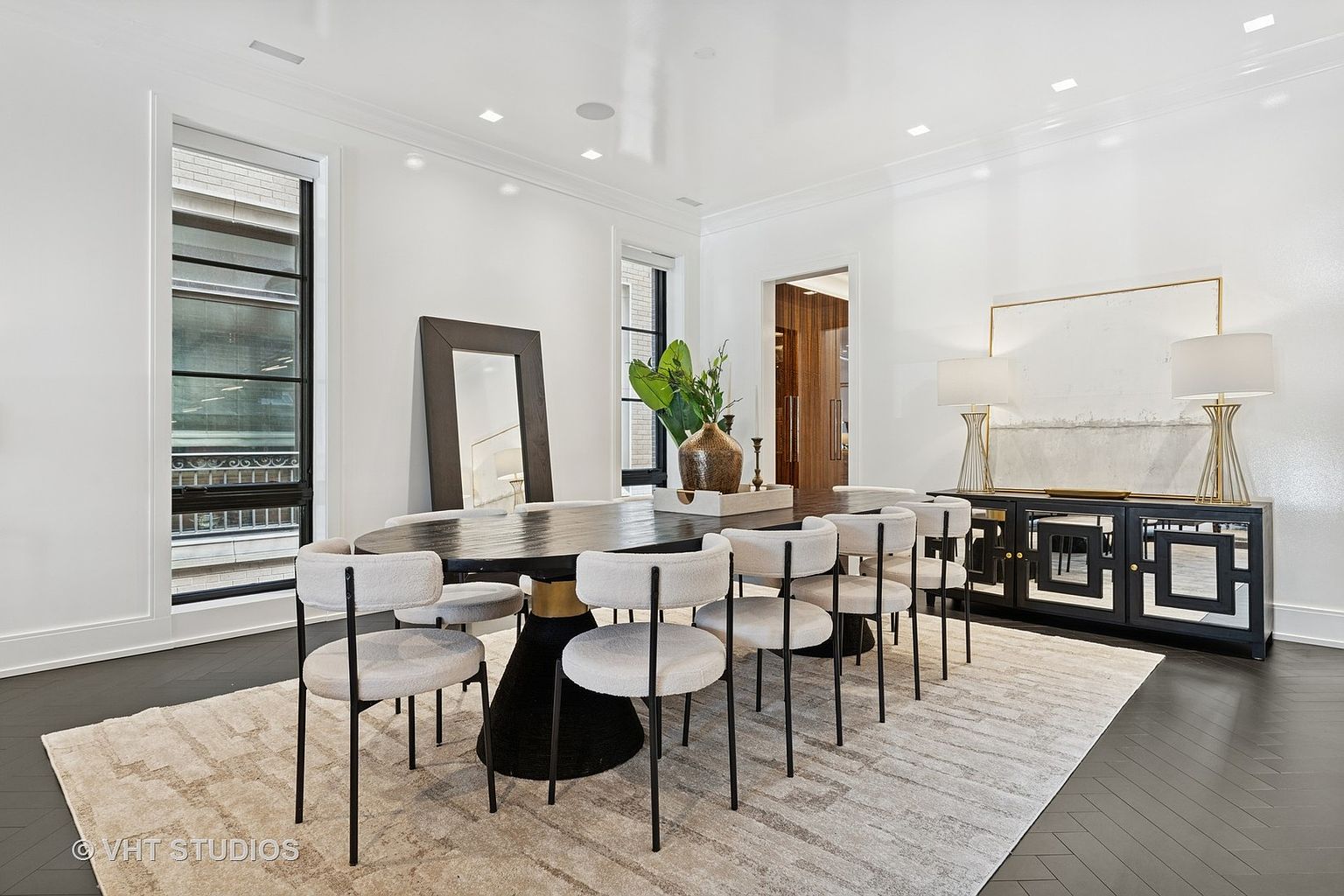 This is an interior shot of a dining room featuring a large oval dining table with a black base and a gold accent, surrounded by eight chairs with light-colored upholstery and black legs. A sideboard with mirrored doors and two lamps sits against the wall, and a large area rug covers the dark hardwood floor. The room is bright and airy, with white walls and trim, creating a sophisticated and elegant atmosphere.