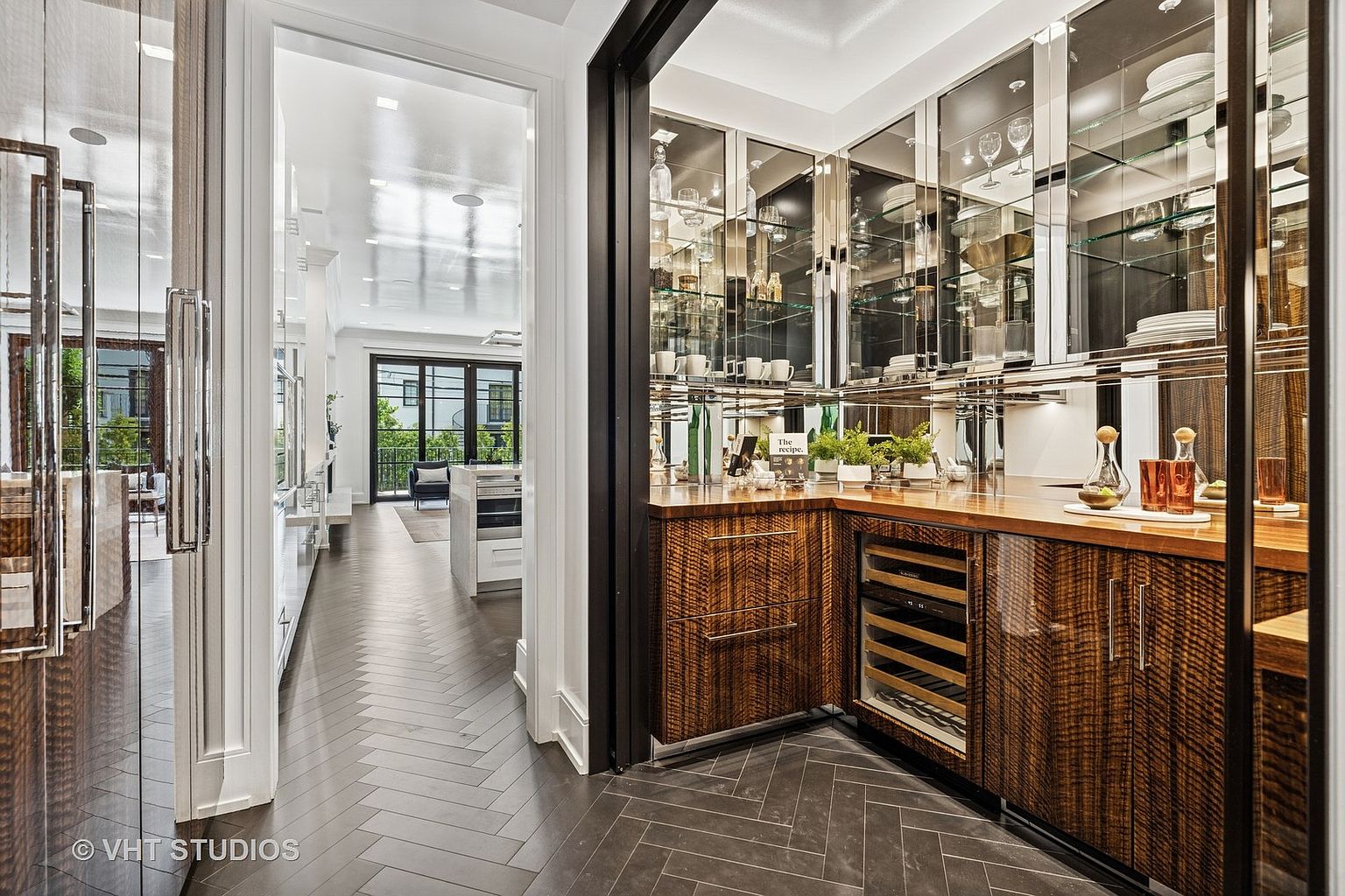 This image showcases a stylish pantry with dark herringbone floors and custom wood cabinetry. Mirrored upper cabinets reflect light and add a touch of elegance, while a built-in wine cooler provides convenience. The pantry is well-organized and designed for both storage and display.