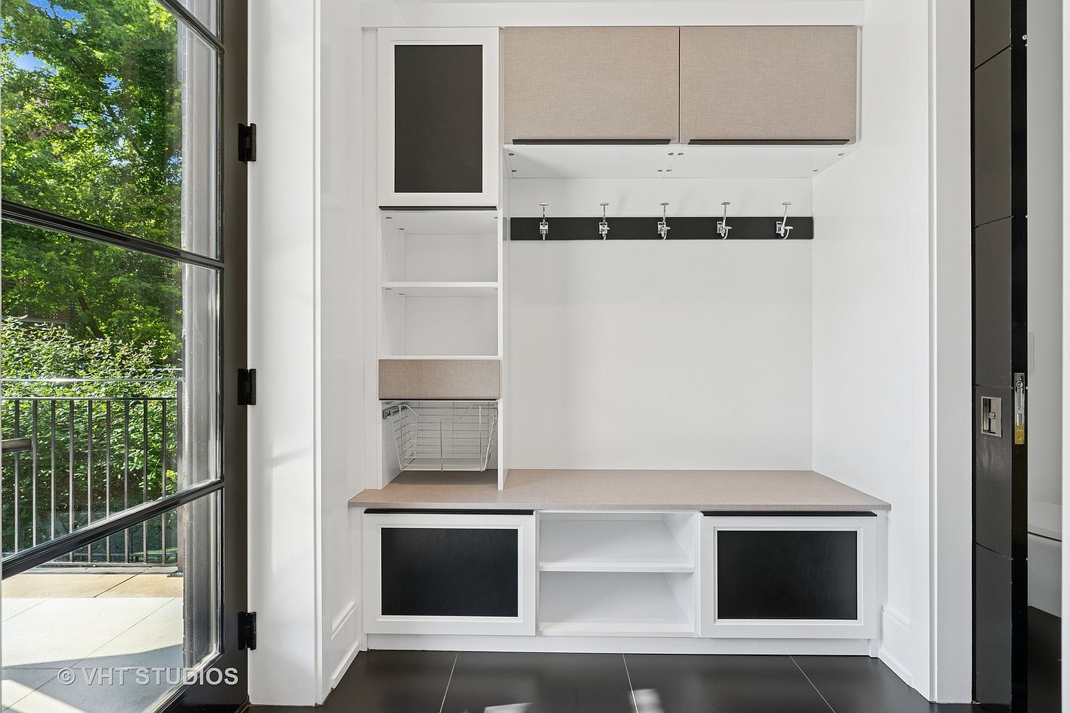 This interior shot showcases a well-organized mudroom area, featuring custom built-in storage with a bench, cabinets, and open shelving. The color palette is primarily white with contrasting black accents, creating a clean and modern aesthetic. The space is designed for functionality and storage, ideal for a busy household.
