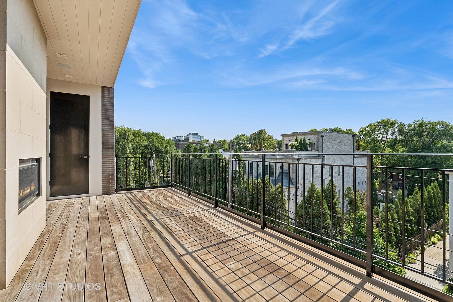 This image showcases a spacious outdoor balcony with a wooden deck and a sleek black metal railing. The view extends over lush greenery and neighboring buildings under a bright blue sky, creating an inviting outdoor living space. A modern fireplace and door are visible on the left, adding architectural interest.
