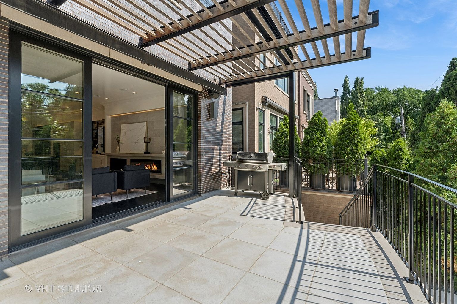 This image showcases a stylish outdoor patio area, featuring a modern pergola that provides partial shade. The patio is furnished with a grill and offers a seamless transition to an indoor living space through large sliding glass doors, revealing a cozy interior with a fireplace. The space is enhanced by a sleek railing and well-maintained landscaping, creating an inviting and sophisticated outdoor living experience.