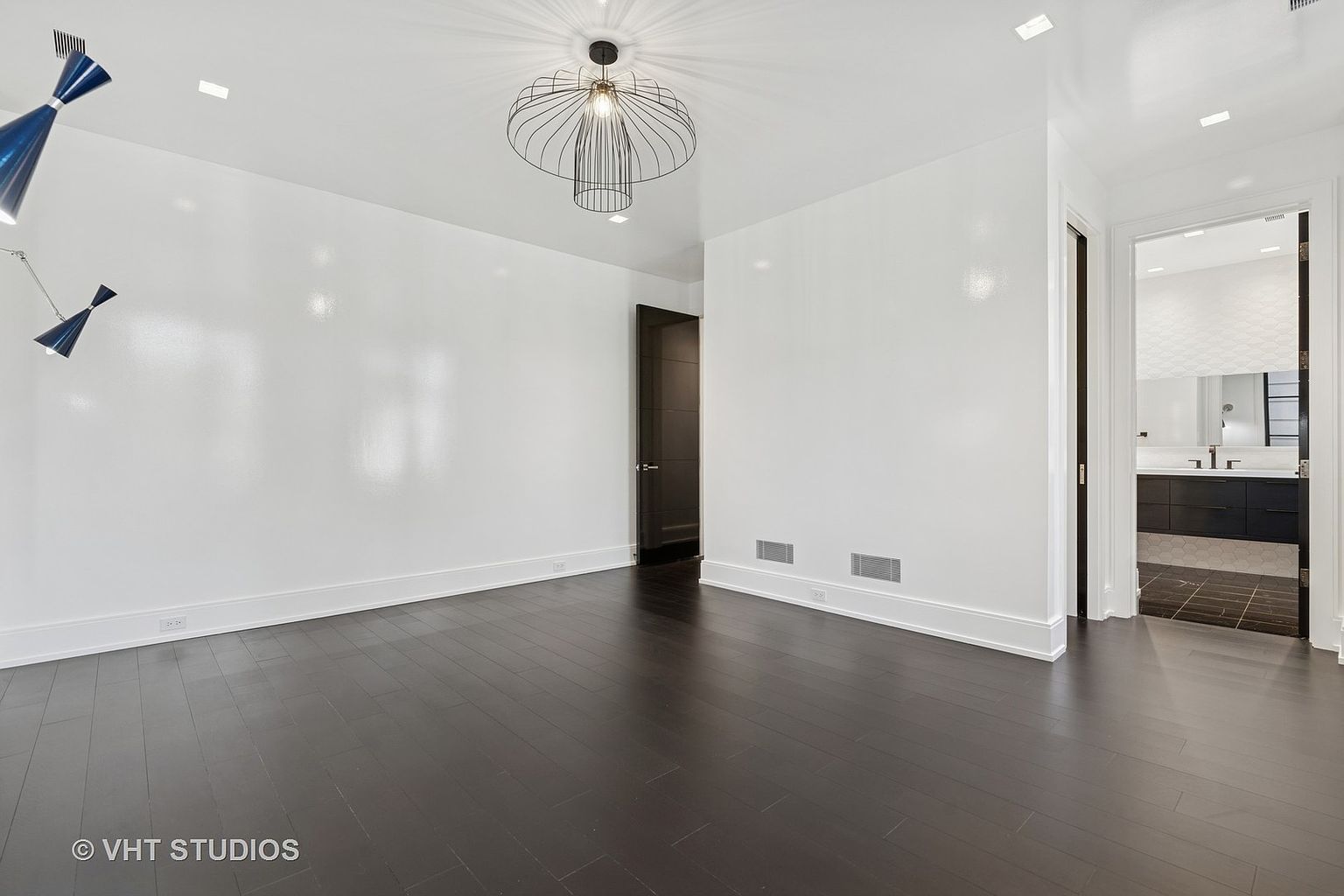 This is a bright and spacious primary bedroom featuring dark hardwood floors and white walls. The room has a modern light fixture and a doorway leading to an en-suite bathroom. The overall impression is clean and minimalist, offering a blank canvas for personalization.