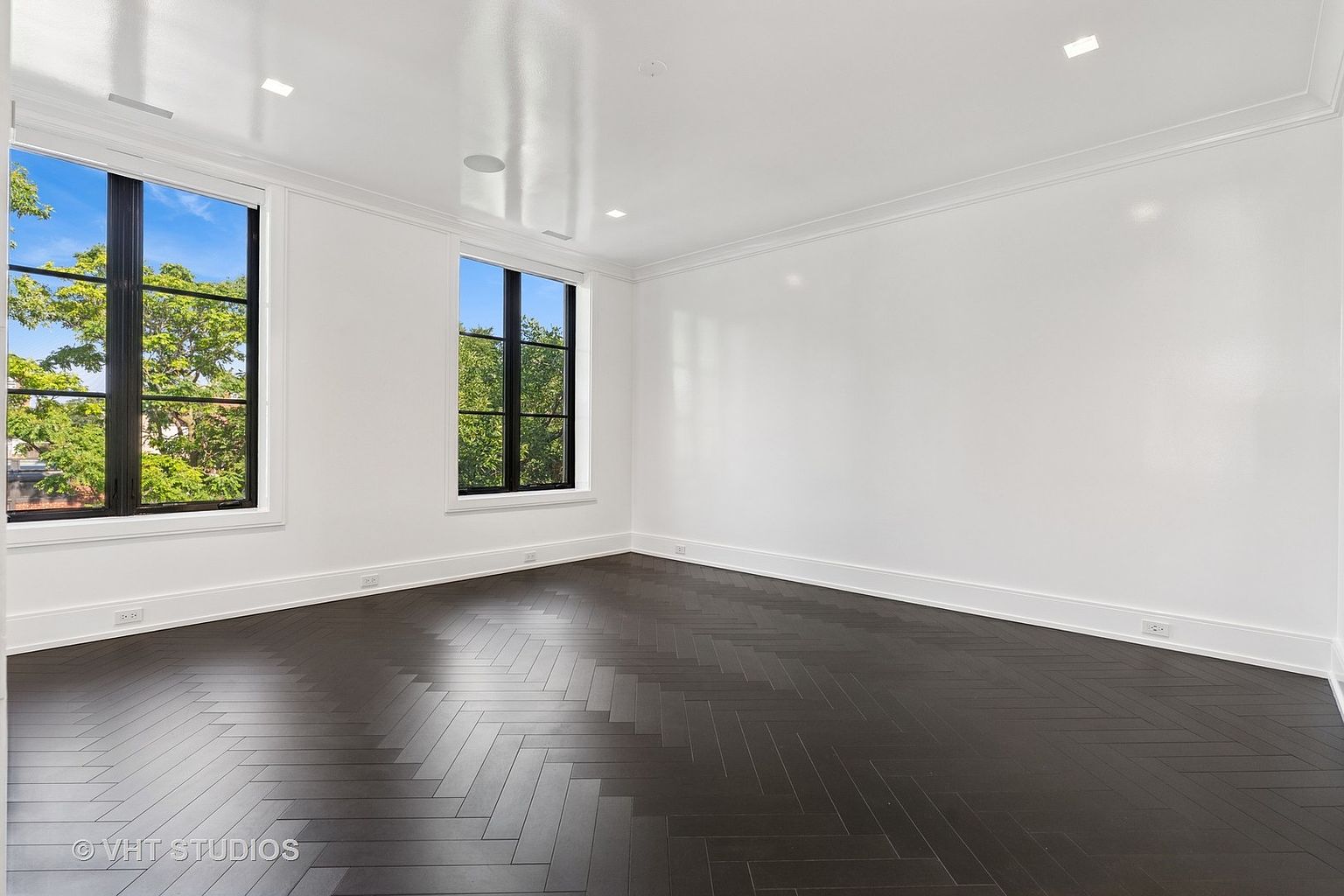 This is a bright, empty room with dark herringbone wood floors, white walls, and white trim. Two windows with black frames let in natural light, and recessed lighting is installed in the ceiling. The room appears clean and modern, ready for customization.