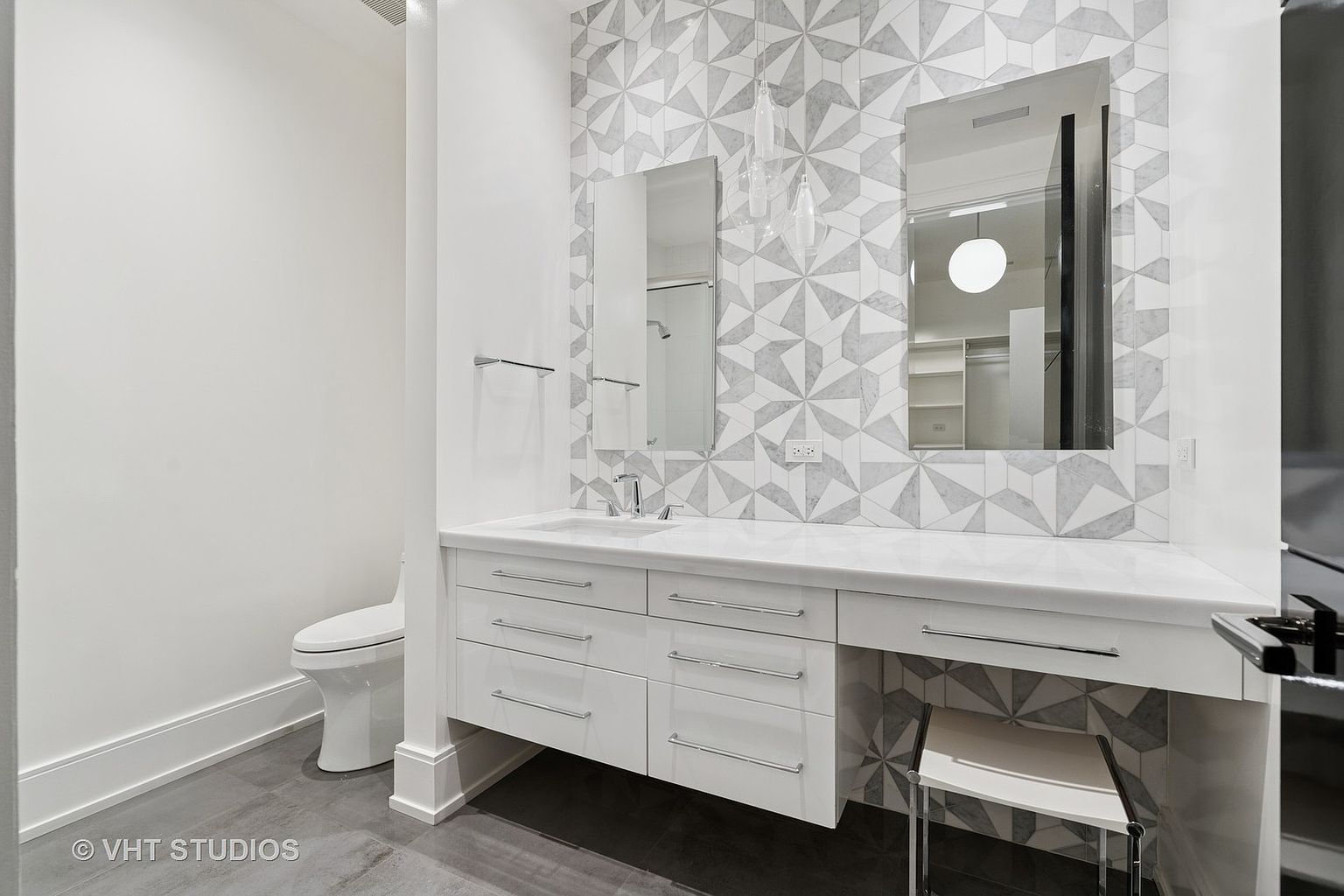 This is a well-lit primary bathroom featuring a modern design with geometric patterned tile backsplash. The vanity is white with sleek, horizontal drawer pulls and a white countertop. Two rectangular mirrors hang above the sink and vanity area, and a toilet is visible to the left. A small stool is tucked under the vanity on the right.