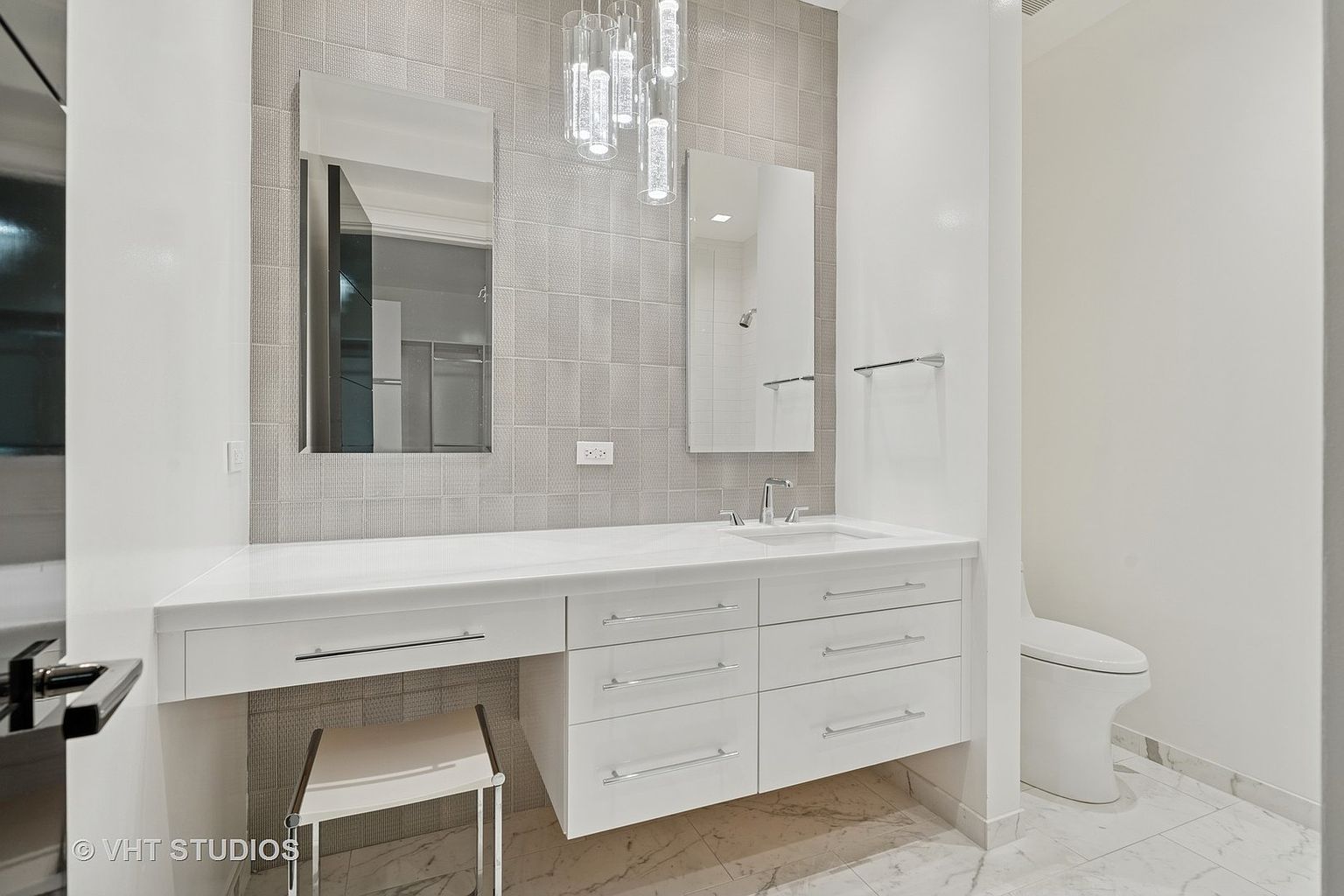 This is a well-lit primary bathroom featuring a modern vanity with white cabinetry and a countertop with a sink. Two rectangular mirrors hang above the vanity, complemented by a decorative pendant light fixture. The walls are tiled in a neutral tone, and a toilet is visible to the right, suggesting a clean and contemporary design.