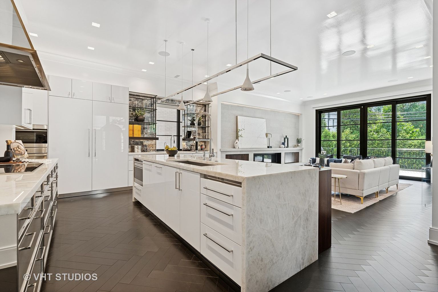 This is a bright, modern kitchen with sleek white cabinetry and a large marble island. The kitchen features high-end appliances and an open layout that flows into the living area, which is visible through large black-framed windows. Dark hardwood floors add a touch of warmth and contrast to the space.
