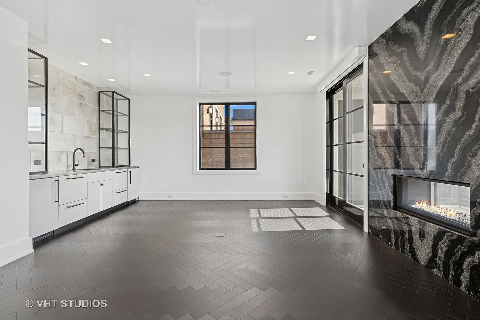 This is a modern living room featuring dark herringbone wood flooring, white walls, and a striking black marble fireplace. A built-in bar area with white cabinetry and glass shelving is on the left, while a window and sliding glass doors provide natural light. The room exudes a sophisticated and contemporary aesthetic.