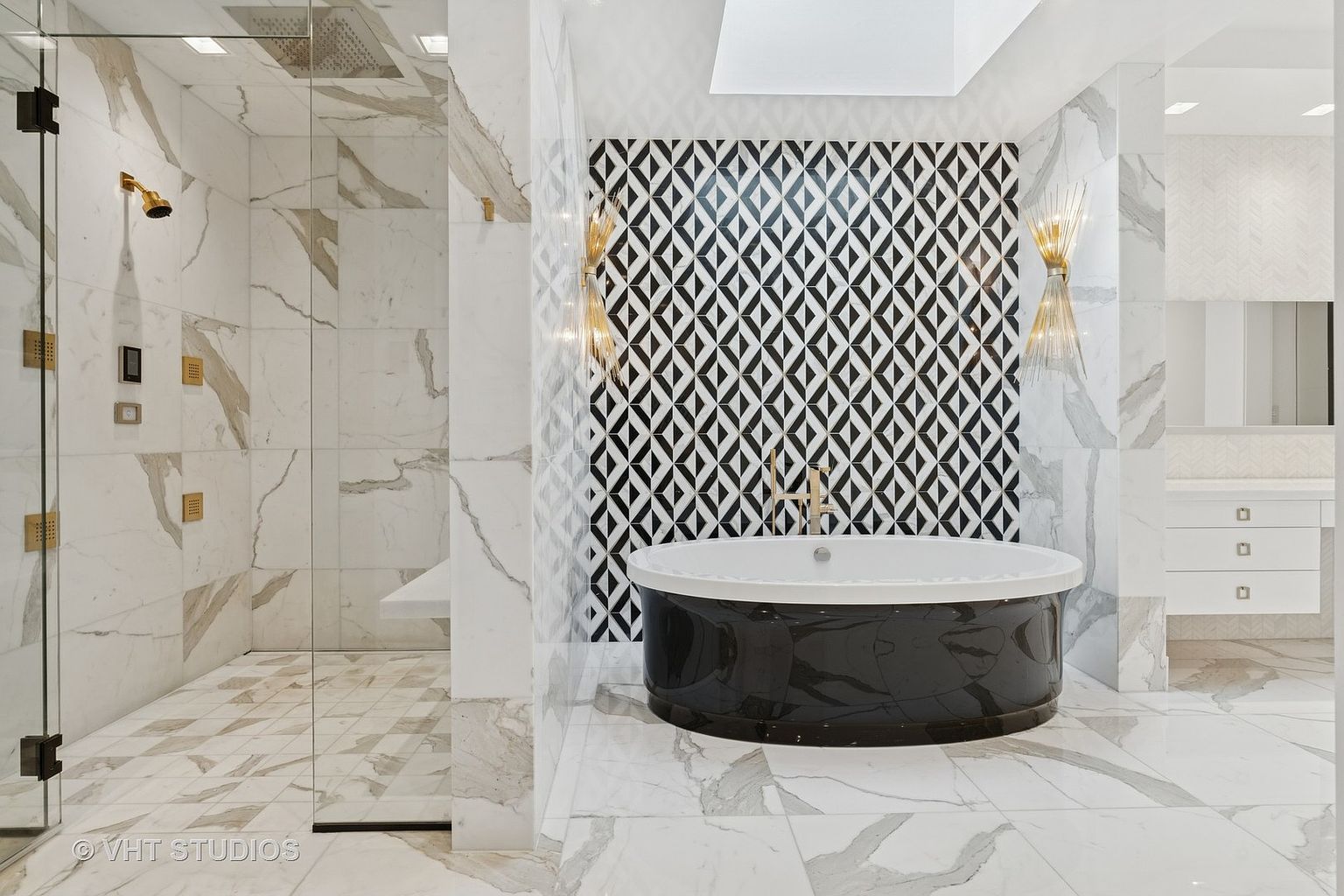 This is a luxurious primary bathroom featuring a modern design with marble walls and floors. A glass-enclosed shower is on the left, while a freestanding black and white bathtub sits against a geometric patterned wall. The bathroom is well-lit and exudes a spa-like atmosphere.