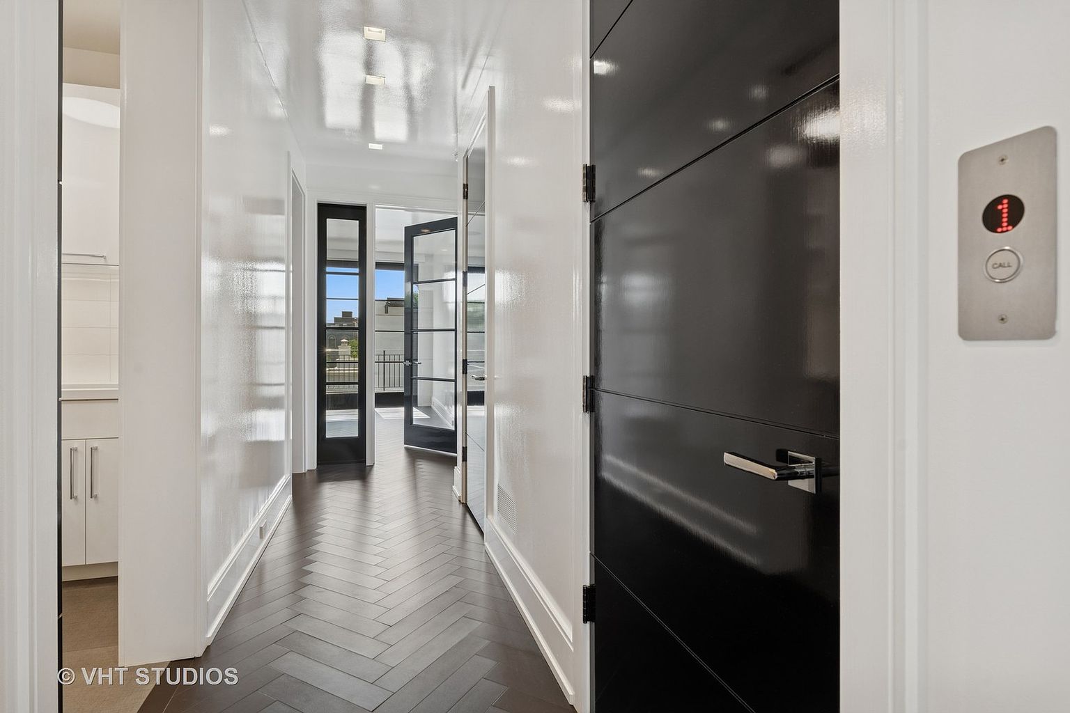 This interior shot showcases a modern hallway with dark herringbone wood flooring and white walls. A black door with horizontal panels is visible on the right, next to a silver elevator call button. The hallway leads to a bright area with black-framed glass doors, suggesting access to an outdoor space or balcony.