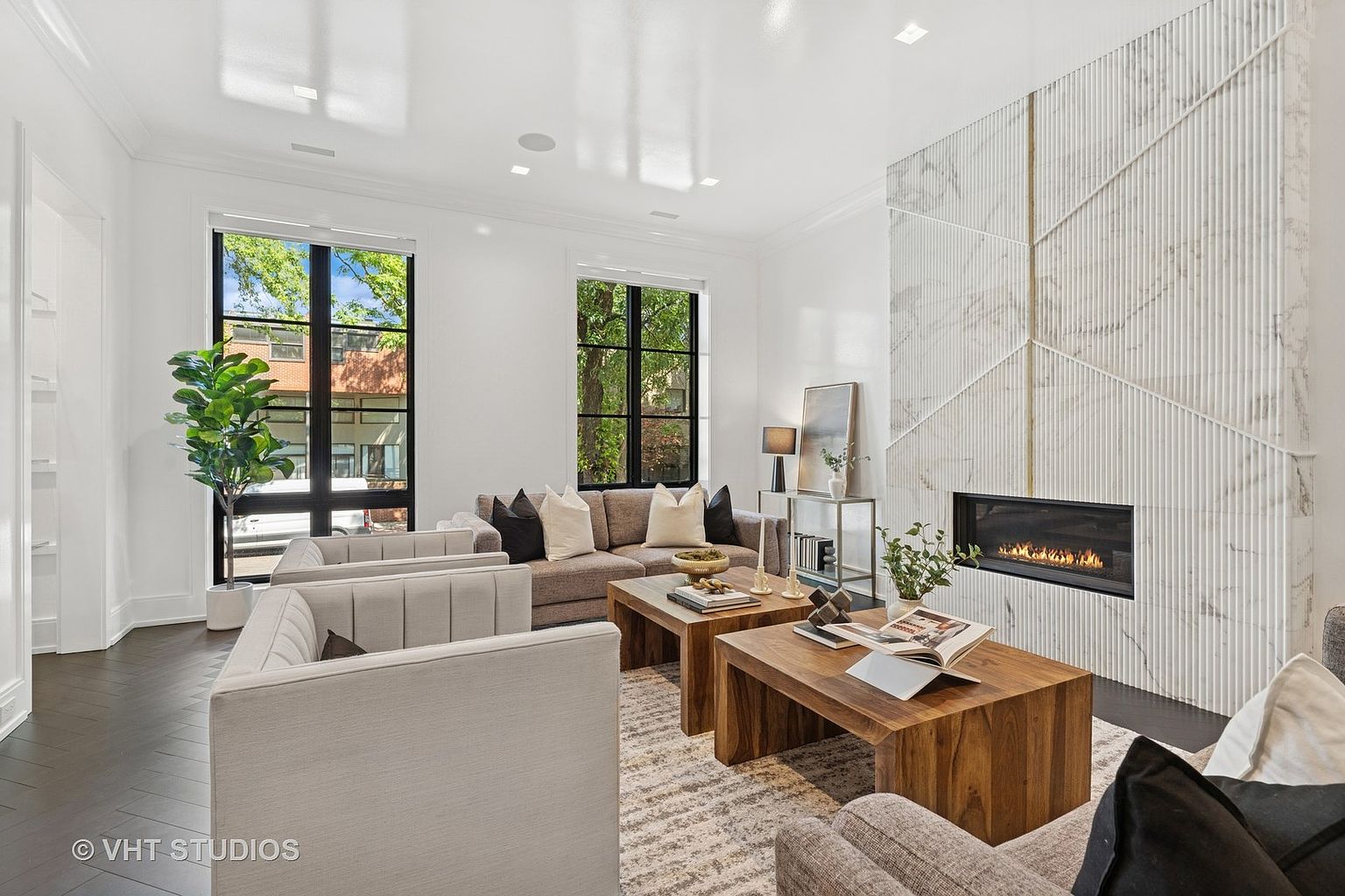 This is a well-lit living room featuring a modern fireplace with a geometric marble design. The room is furnished with a neutral-toned sofa, armchairs, and wooden coffee tables, complemented by a patterned area rug. Large windows provide natural light and views of the outdoors, enhancing the room's bright and airy atmosphere.