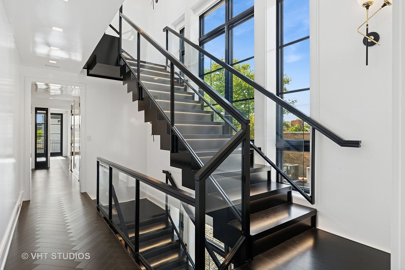 This interior shot showcases a modern staircase with black treads and glass railings, illuminated by subtle lighting. The dark hardwood flooring in a herringbone pattern adds a touch of elegance, while large windows provide ample natural light and a view of the outdoors. The overall impression is clean, contemporary, and sophisticated.