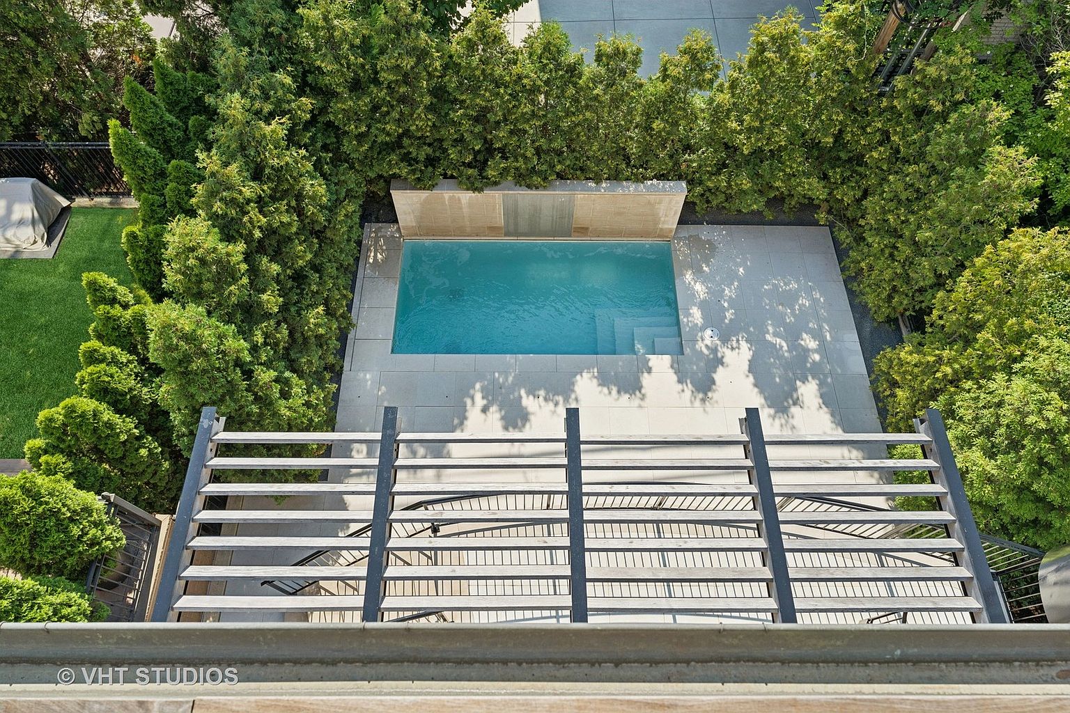 This aerial view showcases a modern rectangular pool surrounded by a light-colored tiled patio and lush green trees, creating a private and serene backyard oasis. A wooden pergola with black metal accents sits in the foreground, adding architectural interest and a sense of enclosure to the outdoor space. The pool's clear blue water invites relaxation and recreation.