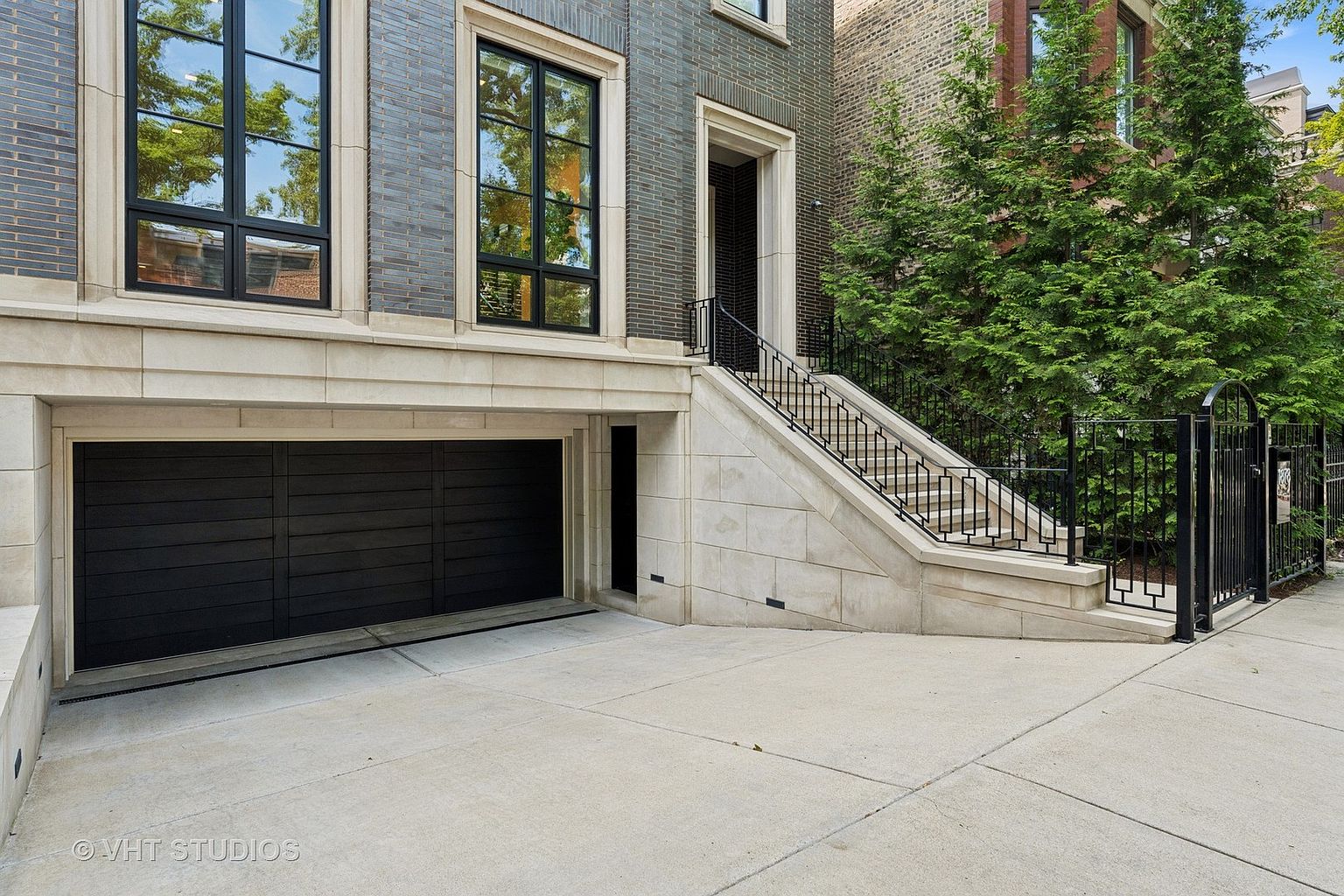 This is a front exterior view of a modern home featuring a dark garage door, a stone facade, and a staircase leading to the main entrance. The property includes well-maintained landscaping with mature trees, adding to its curb appeal. The architectural style is contemporary with clean lines and a sophisticated color palette.