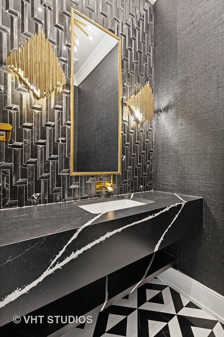 This is a stylish guest bathroom featuring a black marble countertop with white veining and a rectangular sink. The walls are adorned with a mix of textured wallpaper and geometric tile, complemented by gold sconces and a gold-framed mirror. The black and white patterned floor adds a modern touch, creating a sophisticated and luxurious space.