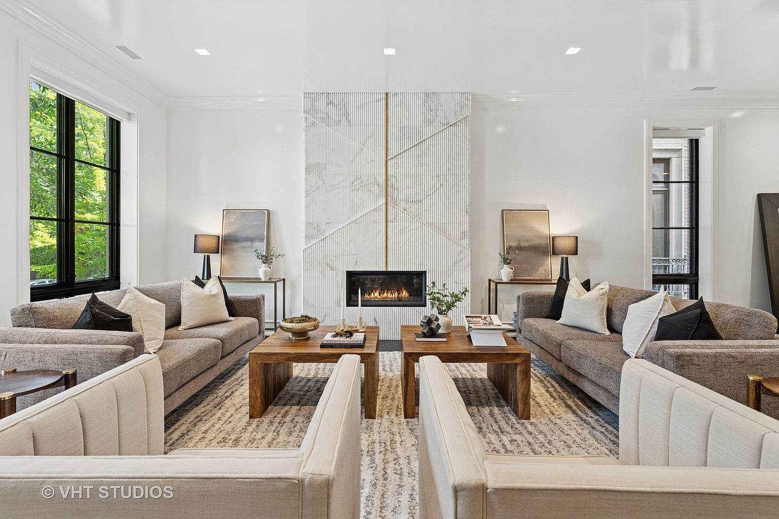 This is a symmetrical living room featuring two sofas facing each other, separated by two wooden coffee tables. A modern fireplace with a geometric marble surround serves as a focal point. The room is bright and airy, with neutral tones and clean lines, creating a sophisticated and inviting atmosphere.