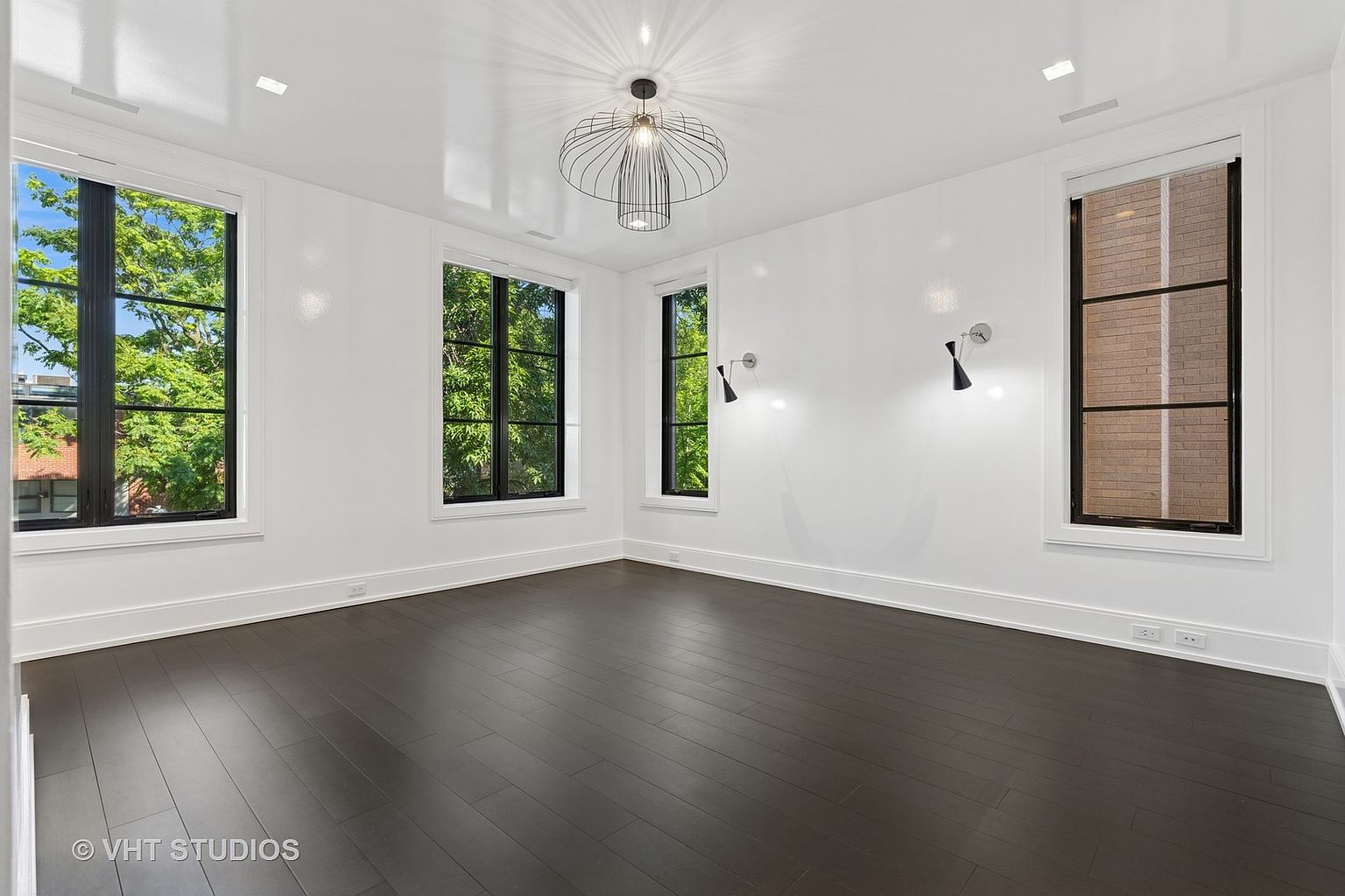 This is a bright and spacious bedroom featuring dark hardwood floors, white walls, and large windows that provide ample natural light. The room is minimally furnished, highlighting the clean lines and modern design. A unique light fixture hangs from the ceiling, adding a touch of contemporary style.