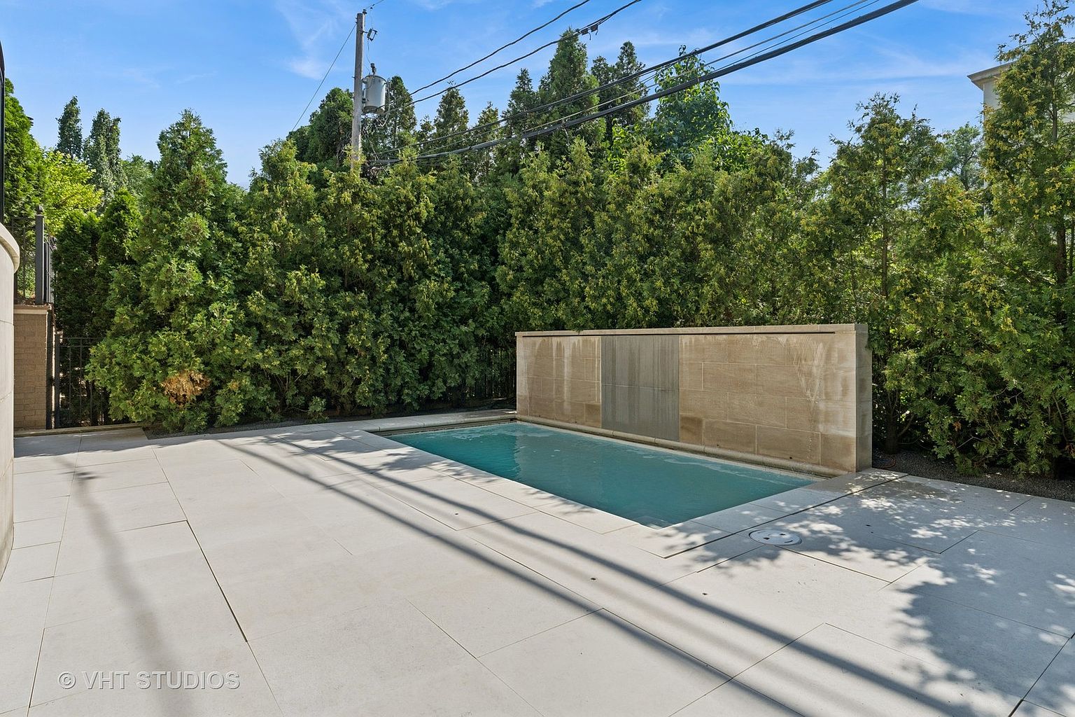 This image showcases a modern pool area, featuring a rectangular pool with a stone waterfall wall as a backdrop. The pool is surrounded by light-colored stone tiling, creating a clean and contemporary aesthetic. Lush green trees and shrubs provide privacy and enhance the serene atmosphere of the outdoor space.