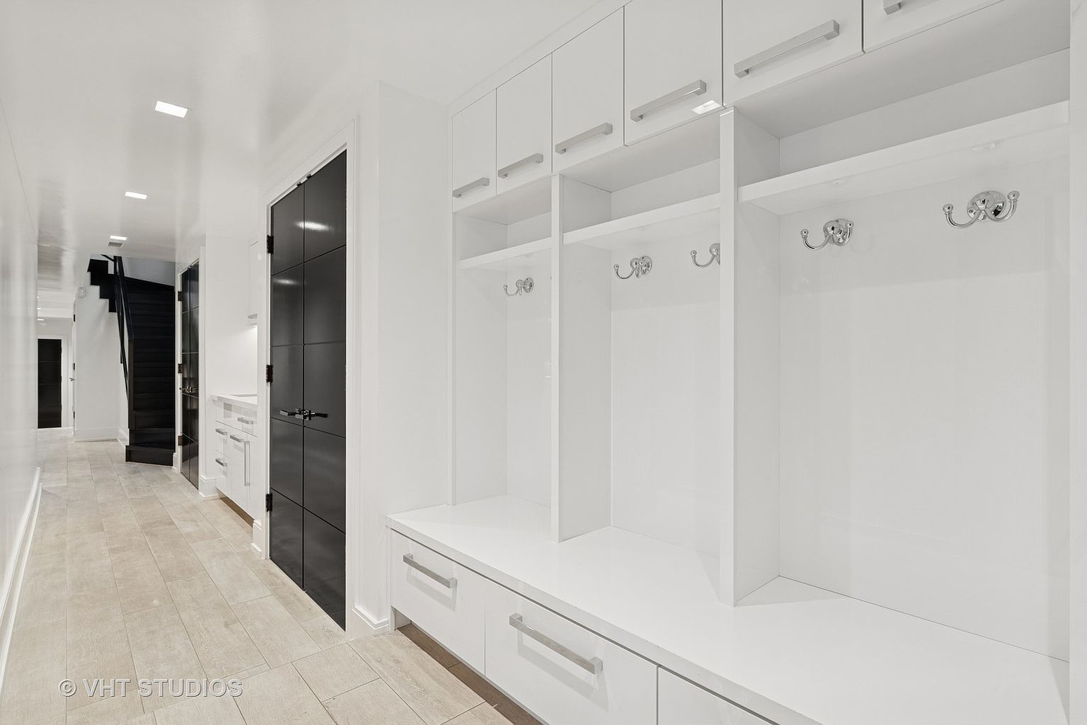 This interior shot showcases a bright hallway with a modern aesthetic, featuring a dark door with a grid pattern and built-in white storage units with drawers and shelving. The hallway leads to a staircase, and the flooring appears to be light-colored tile, creating a clean and spacious feel. The perspective is from the hallway looking towards the stairs.