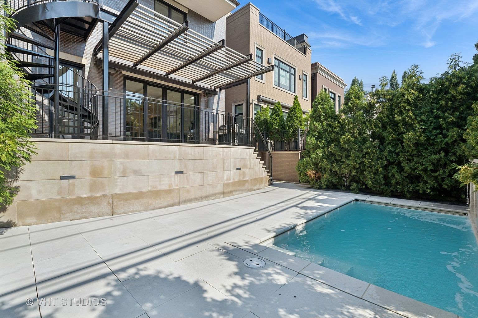 This image showcases the rear exterior of a modern home, highlighting a luxurious pool area. The pool is surrounded by a light-colored patio, and a stone retaining wall supports an elevated deck with a pergola. A spiral staircase adds architectural interest, and mature trees provide privacy.