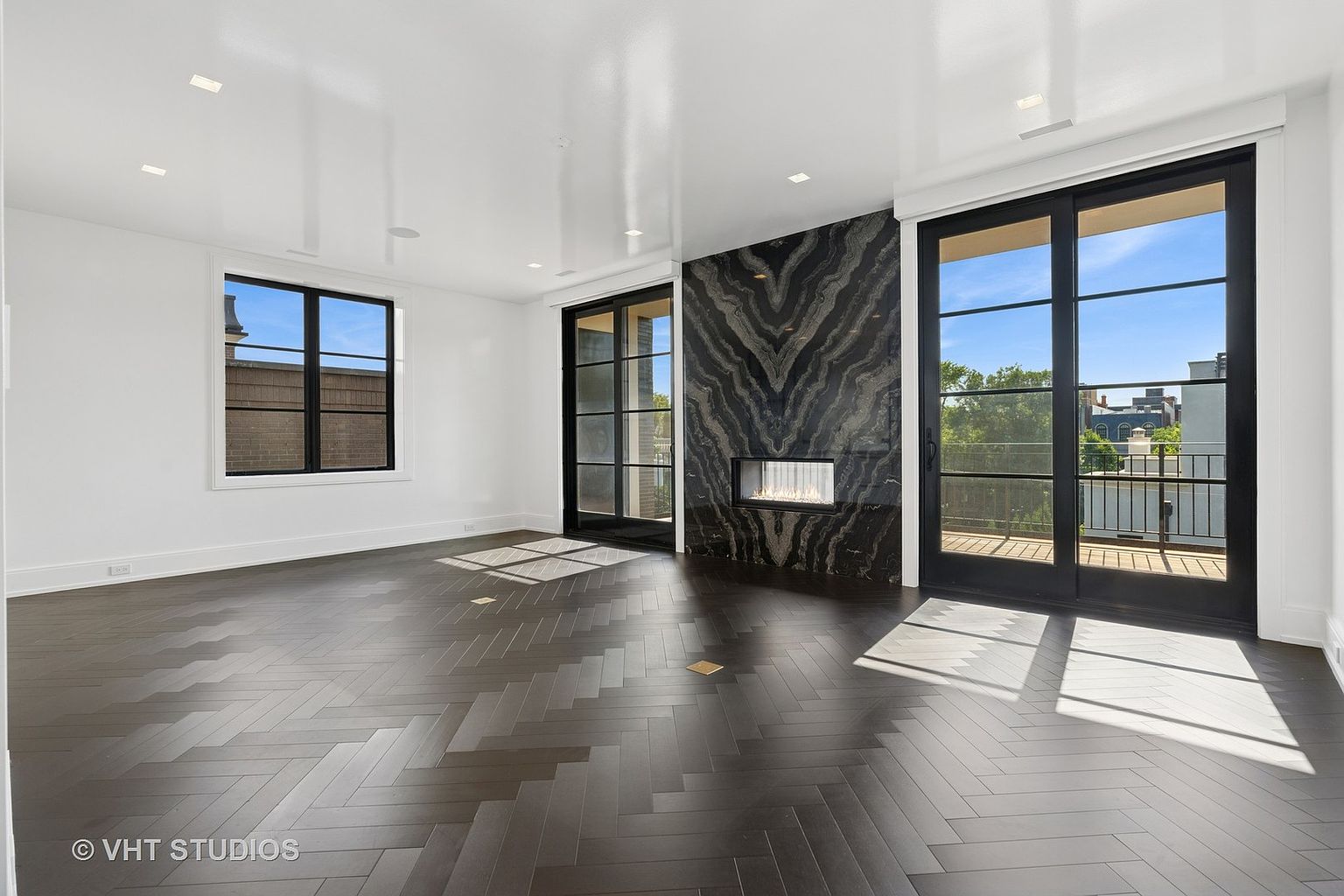 This is a spacious living room featuring dark hardwood floors laid in a herringbone pattern and white walls. A modern fireplace with a striking black and gray marble surround serves as a focal point. Large black-framed windows and sliding glass doors provide ample natural light and access to a balcony, enhancing the room's appeal.
