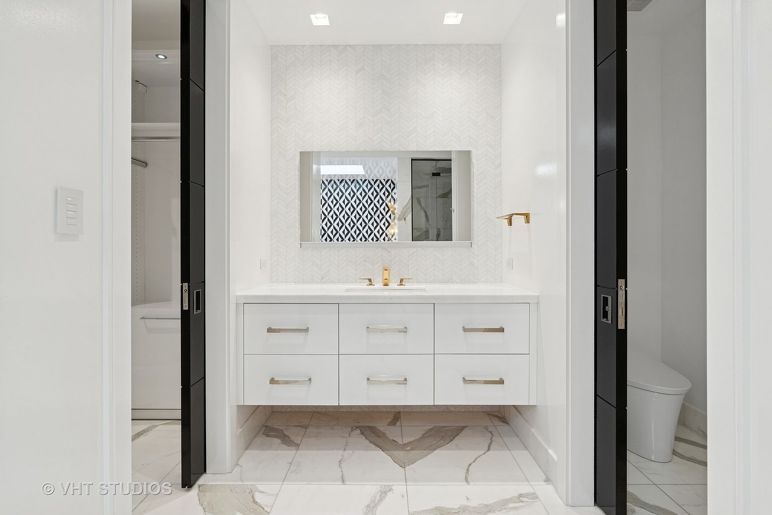 This is a well-lit primary bathroom featuring a floating vanity with white cabinetry and silver hardware, complemented by a white countertop and backsplash with a herringbone pattern. A rectangular mirror hangs above the sink, reflecting a glimpse of another room, and the floor is covered in marble tiles with a unique pattern. The bathroom exudes a clean, modern, and luxurious feel.