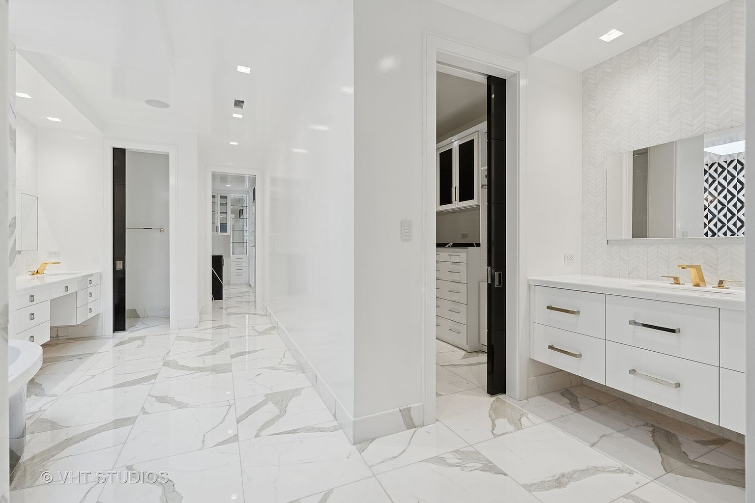 This is a luxurious primary bathroom featuring a pristine white color scheme with marble-like tile flooring. The bathroom includes a double vanity with sleek, modern fixtures and ample storage. The space is well-lit with recessed lighting and natural light, creating a bright and airy atmosphere.