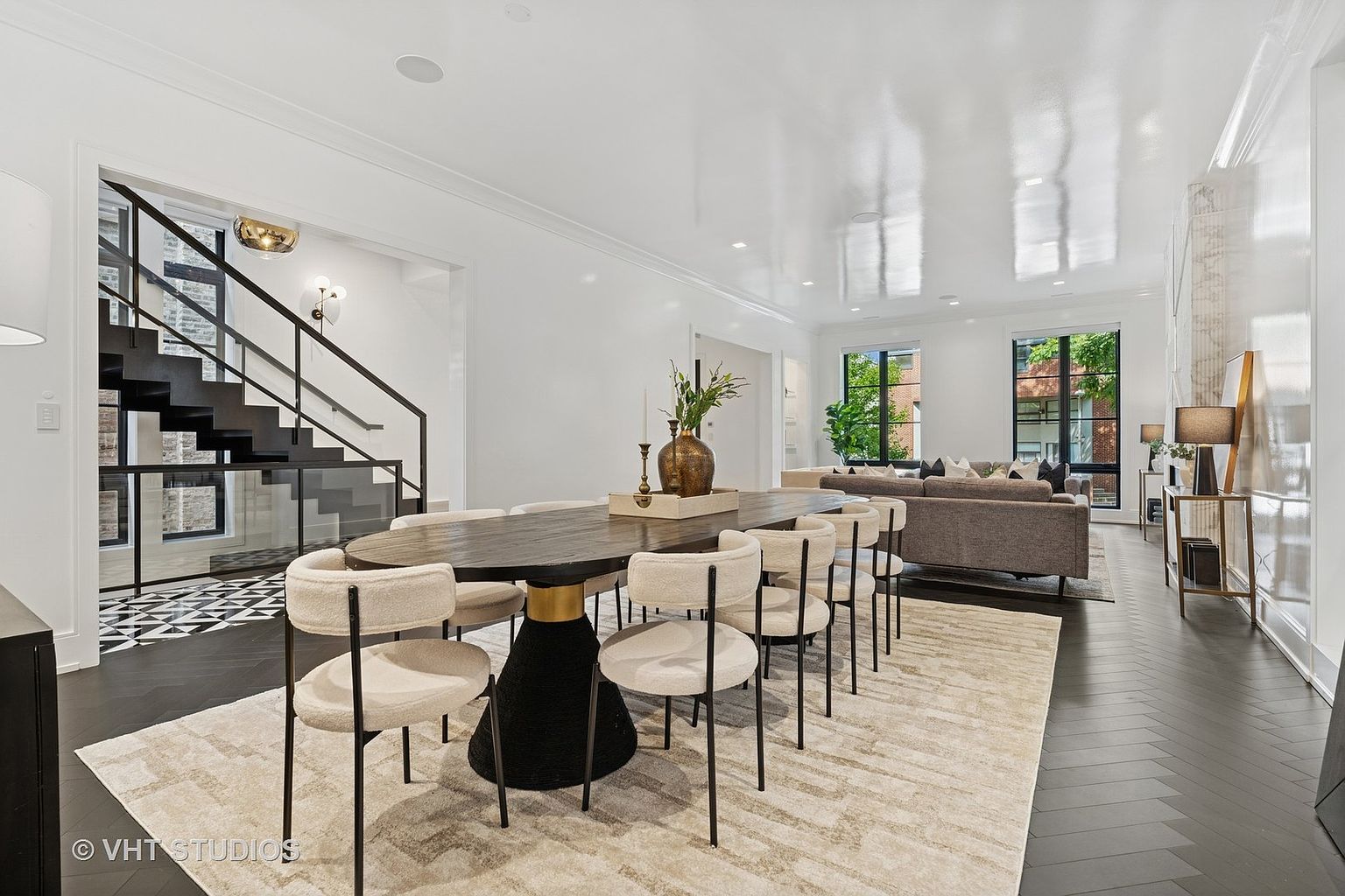 This interior shot showcases a modern dining room with a long, oval-shaped dining table surrounded by stylish chairs with light-colored upholstery and black legs. The room features dark hardwood floors laid in a herringbone pattern, a light-colored area rug beneath the table, and white walls, creating a bright and airy atmosphere that seamlessly transitions into the adjacent living area. A staircase with black railings is visible in the background, adding architectural interest.