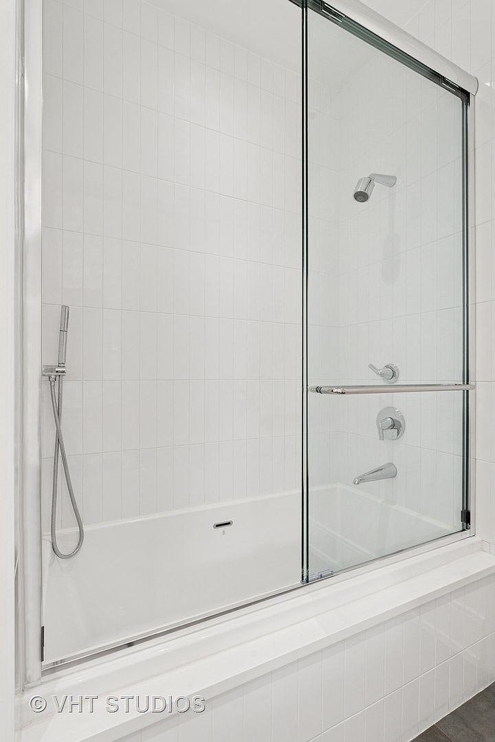 This is a well-lit bathroom featuring a bathtub with a glass shower door. The walls are tiled in a clean, white subway style, and the fixtures are chrome. The overall impression is modern and minimalist, emphasizing cleanliness and functionality.