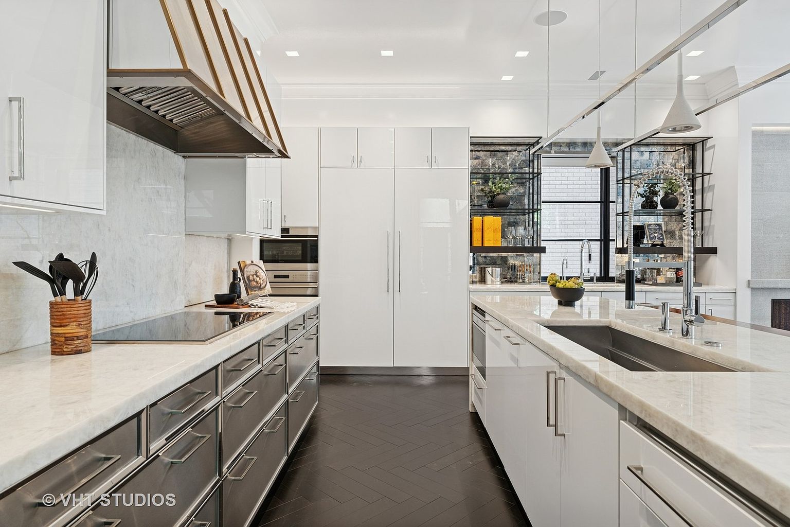 This is a modern kitchen with sleek white cabinetry and stainless steel accents. The kitchen features a large island with a marble countertop and a stainless steel sink. The dark wood flooring adds a touch of warmth to the space, and the open shelving provides a display area for decorative items.