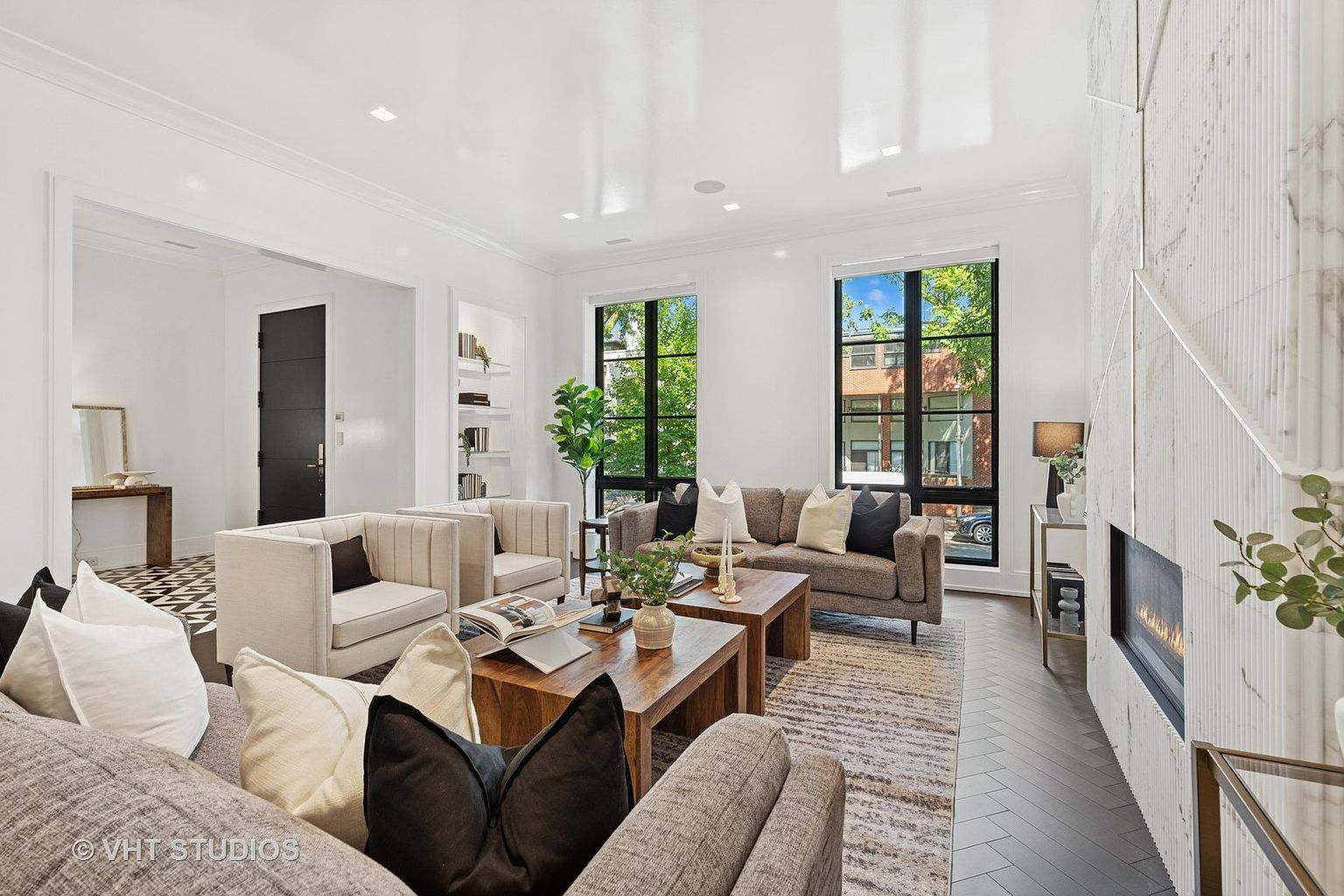 This is an interior shot of a modern living room featuring neutral tones and clean lines. The room is well-lit with natural light streaming through large windows, highlighting the hardwood floors and contemporary furniture. A fireplace with a unique marble surround adds a focal point, while the overall design creates a sophisticated and inviting atmosphere.