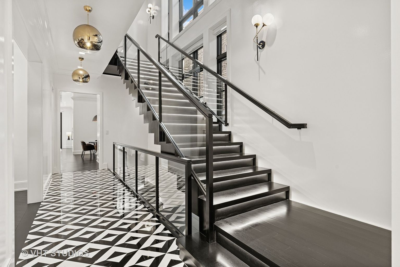 This interior shot showcases a modern hallway and staircase. The staircase features dark wood steps with integrated lighting and glass railings, complemented by a black handrail. The hallway boasts a striking black and white geometric tile floor, leading to a glimpse of a dining area, while gold pendant lights add a touch of elegance.