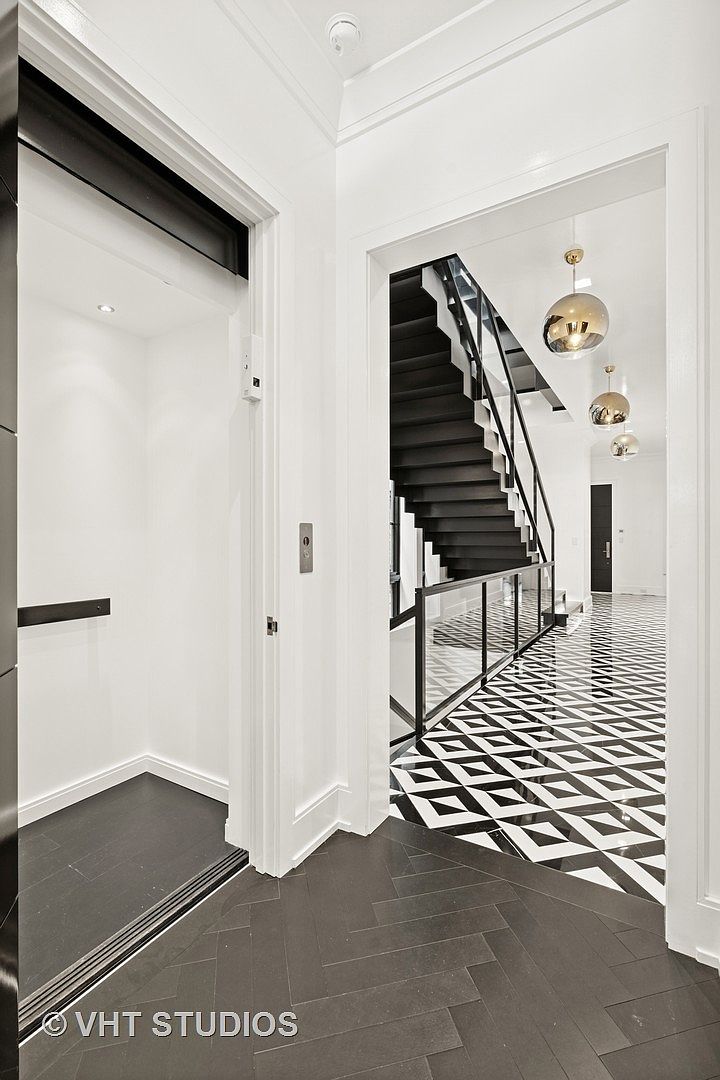 This interior shot showcases a modern hallway with striking black and white geometric tile flooring. A black staircase with glass railings is visible through an archway, complemented by elegant gold pendant lights. To the left, an elevator adds a touch of luxury and convenience, while the dark wood flooring in the elevator area contrasts with the hallway's tile.