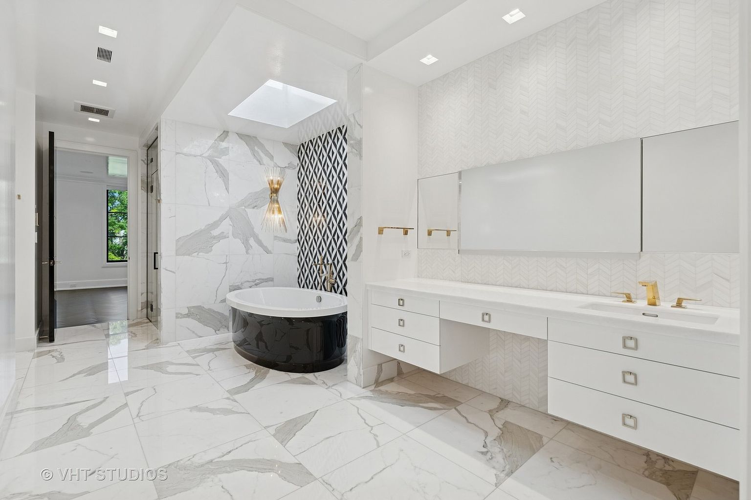 This is a luxurious primary bathroom featuring a modern design with white marble flooring and walls accented by a black and white geometric patterned wall. A freestanding black soaking tub sits beneath a skylight, while a long white vanity with gold fixtures and a large mirror spans the opposite wall. The space is bright and airy, creating a spa-like atmosphere.