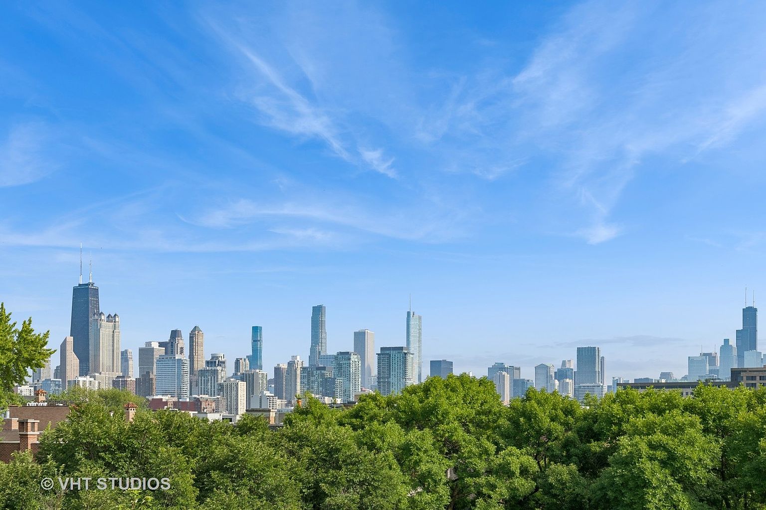 This aerial view showcases a stunning cityscape under a bright blue sky with wispy clouds. Lush green trees fill the foreground, providing a natural contrast to the towering buildings in the distance. The image captures the scale and beauty of the urban environment, highlighting the proximity of nature to the city.