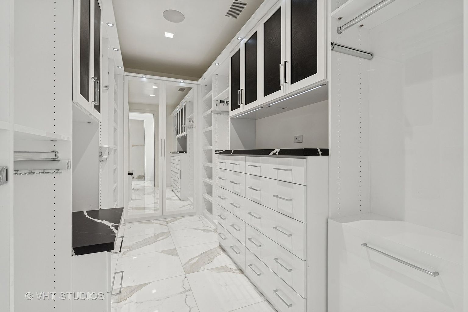 This is a well-organized walk-in closet featuring custom shelving, drawers, and hanging rods. The closet is brightly lit and has a clean, modern aesthetic with white cabinetry and marble flooring. The design maximizes storage space and provides a luxurious feel.