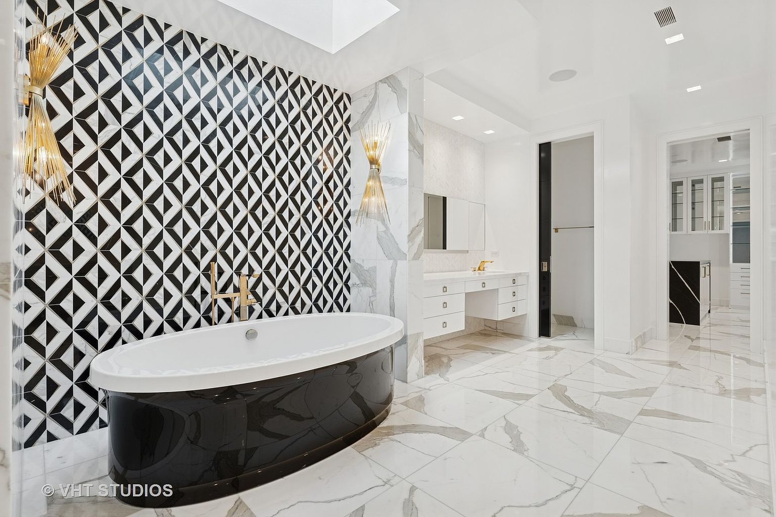 This is a luxurious primary bathroom featuring a freestanding black and white bathtub against a striking black and white geometric tiled wall. The bathroom boasts marble flooring, a white vanity with gold fixtures, and elegant lighting. The space is bright and airy, creating a spa-like atmosphere.
