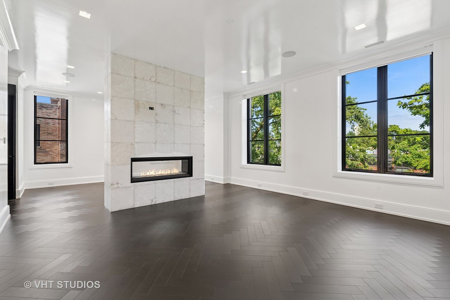 This is a bright and spacious living room featuring dark herringbone wood flooring and white walls. A modern fireplace with a light marble surround serves as a focal point, and large windows with black frames offer views of the outdoors. The room exudes a clean and contemporary aesthetic, perfect for showcasing the home's modern design.