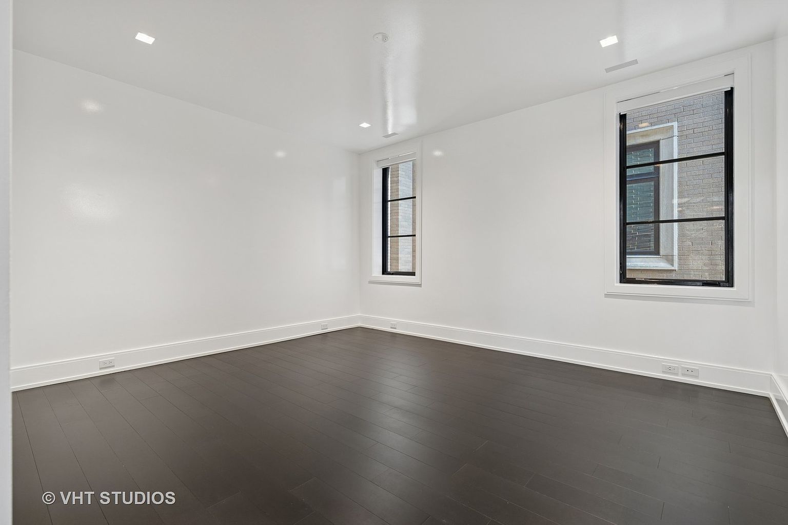 This is a bright, minimalist bedroom featuring dark hardwood floors and white walls. Two windows with black frames provide natural light and views of the exterior. The room is clean and uncluttered, offering a blank canvas for potential buyers to envision their own style.