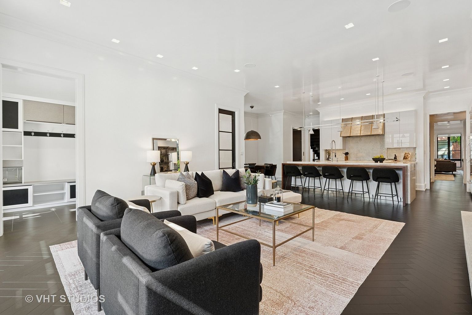 This is an interior shot of a modern living room that seamlessly flows into the kitchen. The living room features a neutral color palette with dark hardwood floors, a light-colored sectional sofa, and a glass coffee table. The kitchen has a large island with bar seating and modern cabinetry, creating an open and inviting space perfect for entertaining.