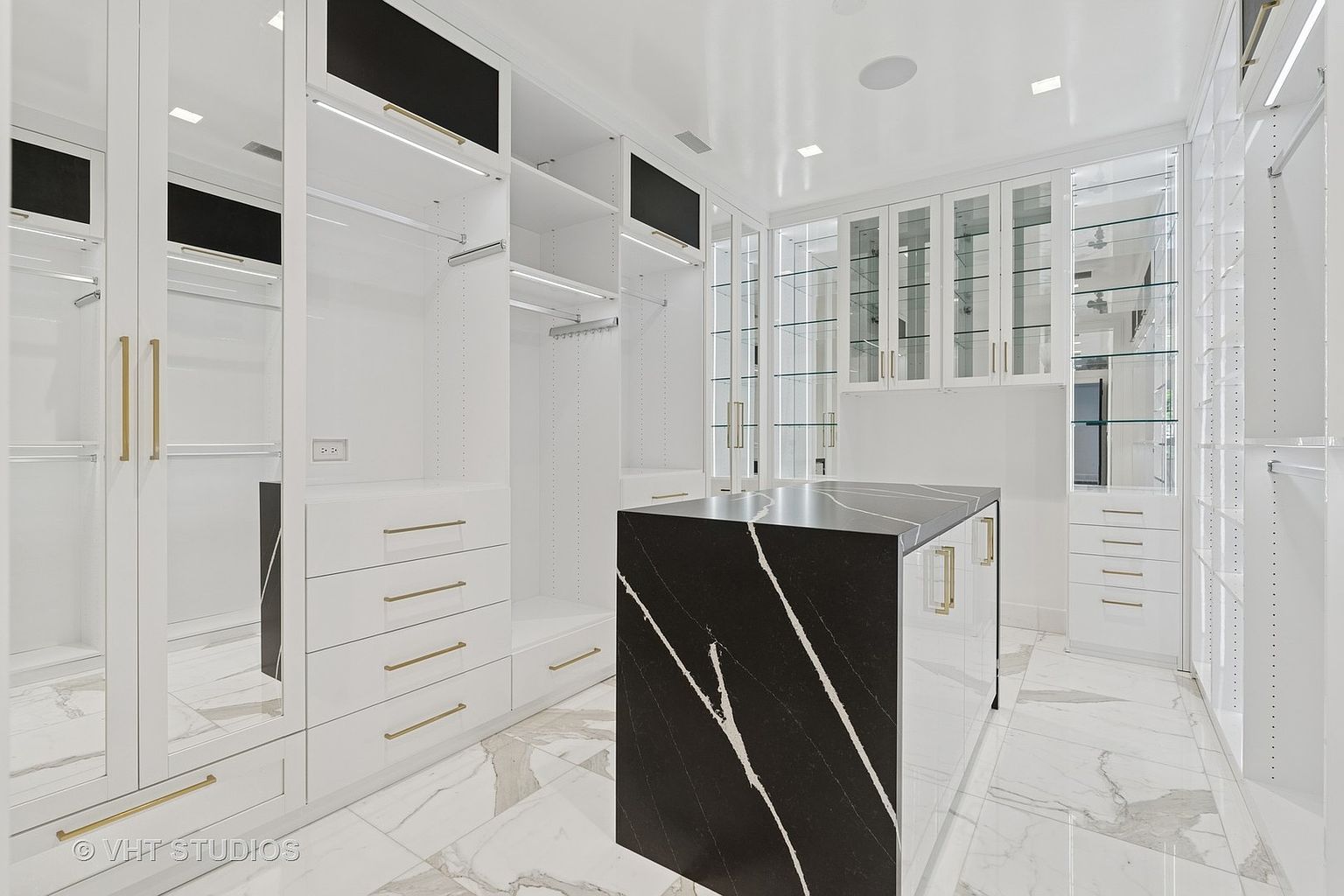This is a luxurious walk-in closet featuring custom white cabinetry with gold hardware and a black marble island. The closet is well-lit with recessed lighting and features mirrored doors, drawers, shelves, and hanging rods for ample storage. The marble flooring adds to the high-end aesthetic, creating a sophisticated and organized space.