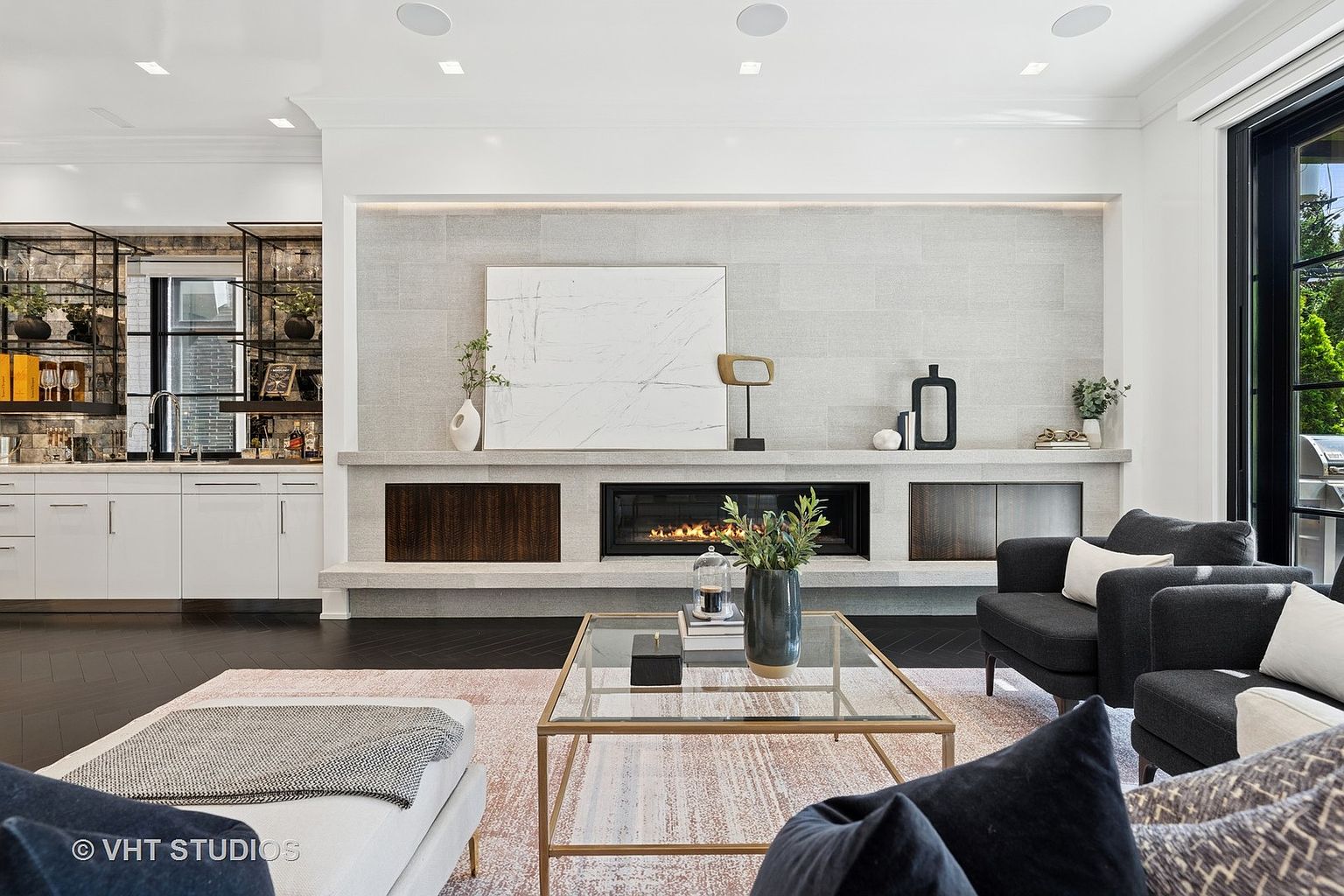 This is an interior shot of a modern living room featuring a fireplace with a light gray stone surround and dark wood accents. The room is furnished with dark gray armchairs, a glass-topped coffee table, and a light-colored sofa with a patterned throw. A built-in bar area with open shelving is visible in the background, adding to the room's sophisticated and inviting atmosphere.