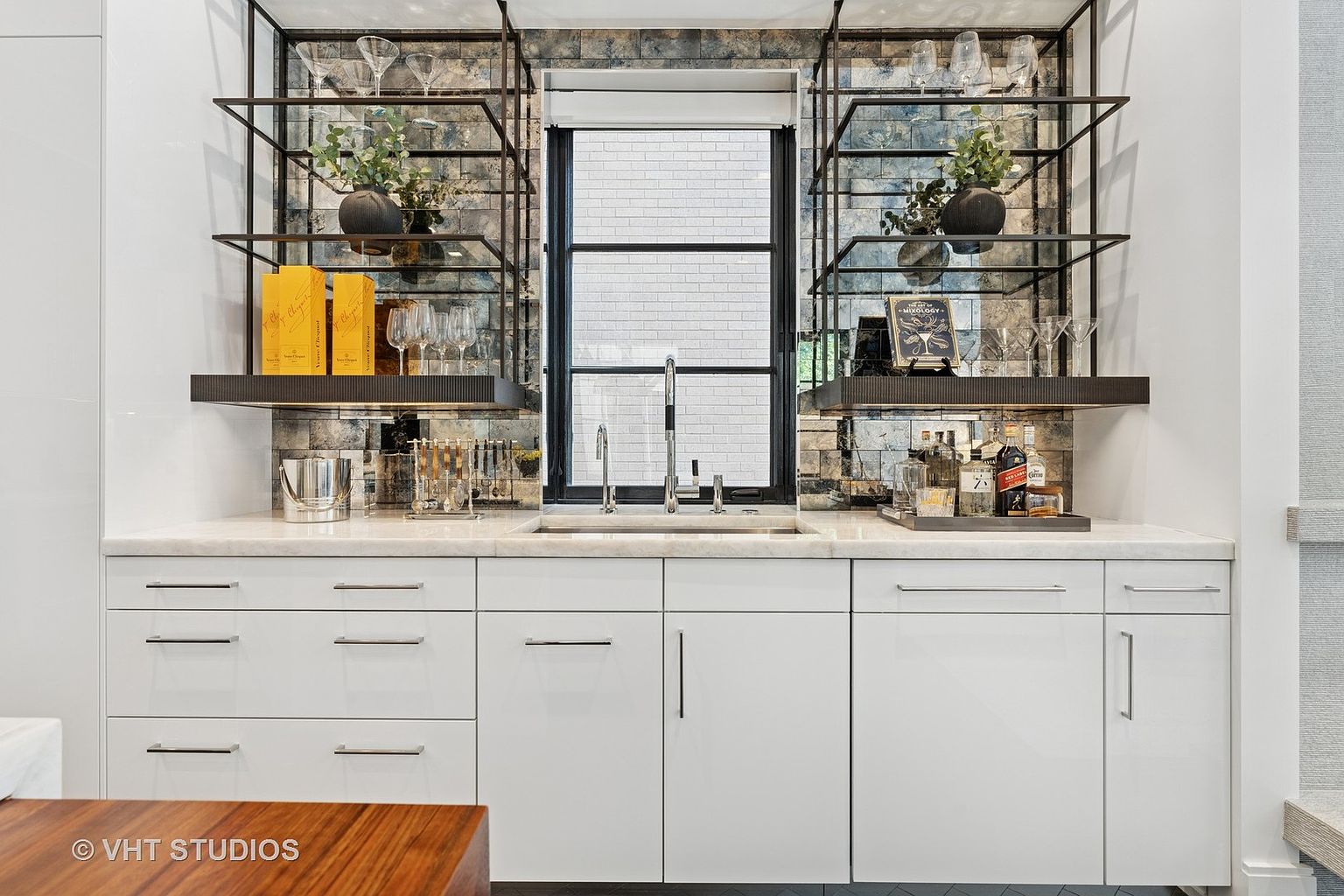 This interior shot showcases a stylish kitchen bar area with white cabinetry, a marble countertop, and open shelving displaying glassware and decorative items. A window above the sink provides natural light, and the backsplash features a unique mirrored tile design. The overall aesthetic is modern and sophisticated, perfect for entertaining.
