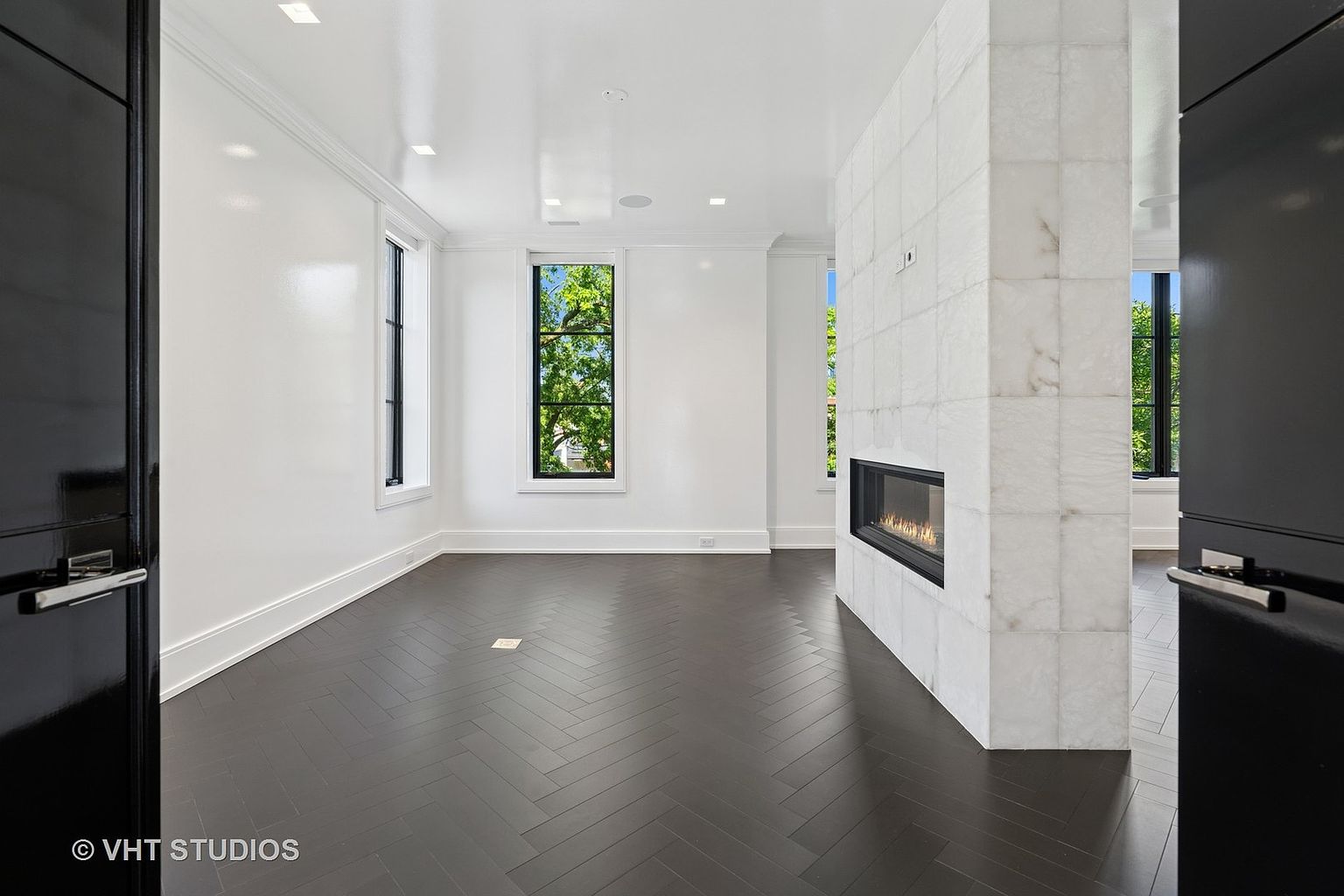 This is an interior shot of a modern living room featuring dark herringbone hardwood floors and white walls. A striking marble fireplace divides the space, adding a focal point. Natural light streams in through the windows, creating a bright and airy atmosphere.
