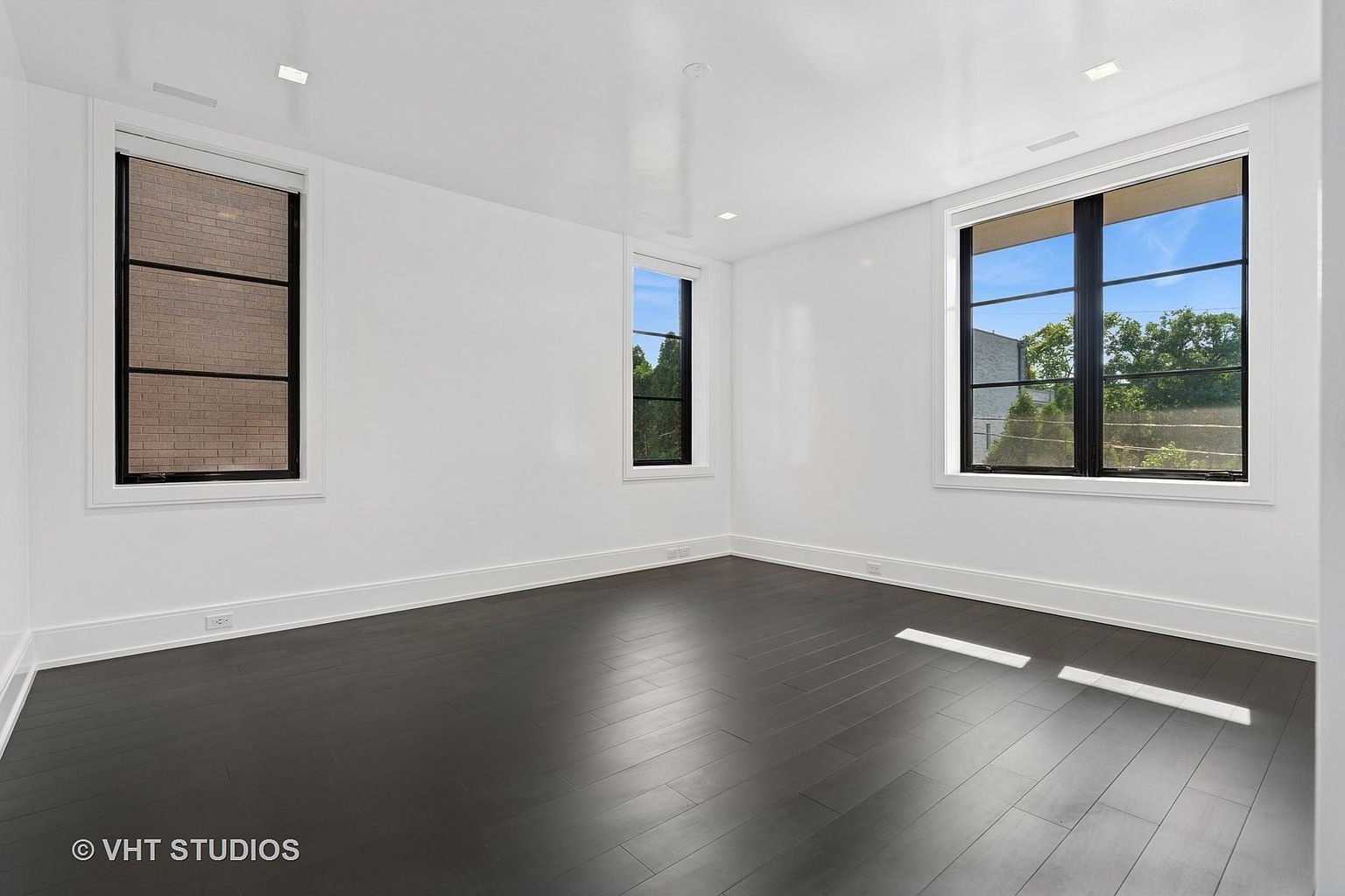 This is a bright and modern bedroom featuring dark hardwood floors and white walls. Three windows provide natural light, with one offering a view of a brick building and another showcasing greenery. The room is clean and minimalist, creating a spacious and airy feel.