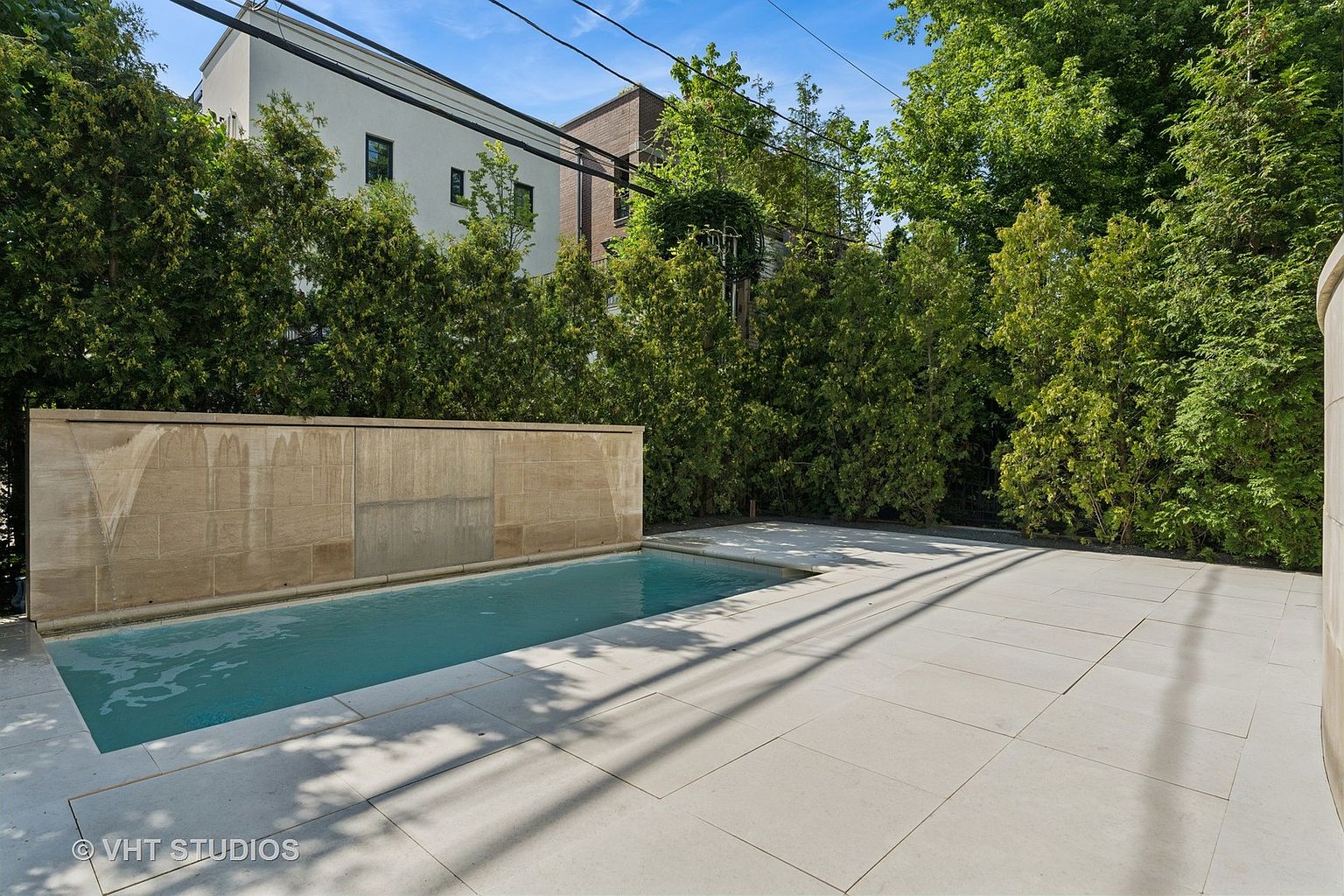 This image showcases a modern outdoor pool area with a sleek, minimalist design. The pool is surrounded by light-colored stone paving, and a textured stone wall serves as a backdrop, possibly incorporating a water feature. Lush greenery provides privacy and a serene atmosphere, enhancing the property's appeal for relaxation and entertainment.