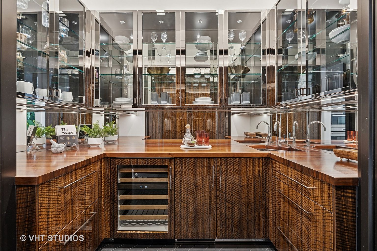 This is an interior shot of a modern kitchen featuring wood cabinetry with a unique chevron pattern and a rich, warm tone. Mirrored upper cabinets display glassware and dishware, while a wine refrigerator is integrated into the lower cabinetry. The countertops are a polished wood, and the overall design exudes luxury and sophistication.
